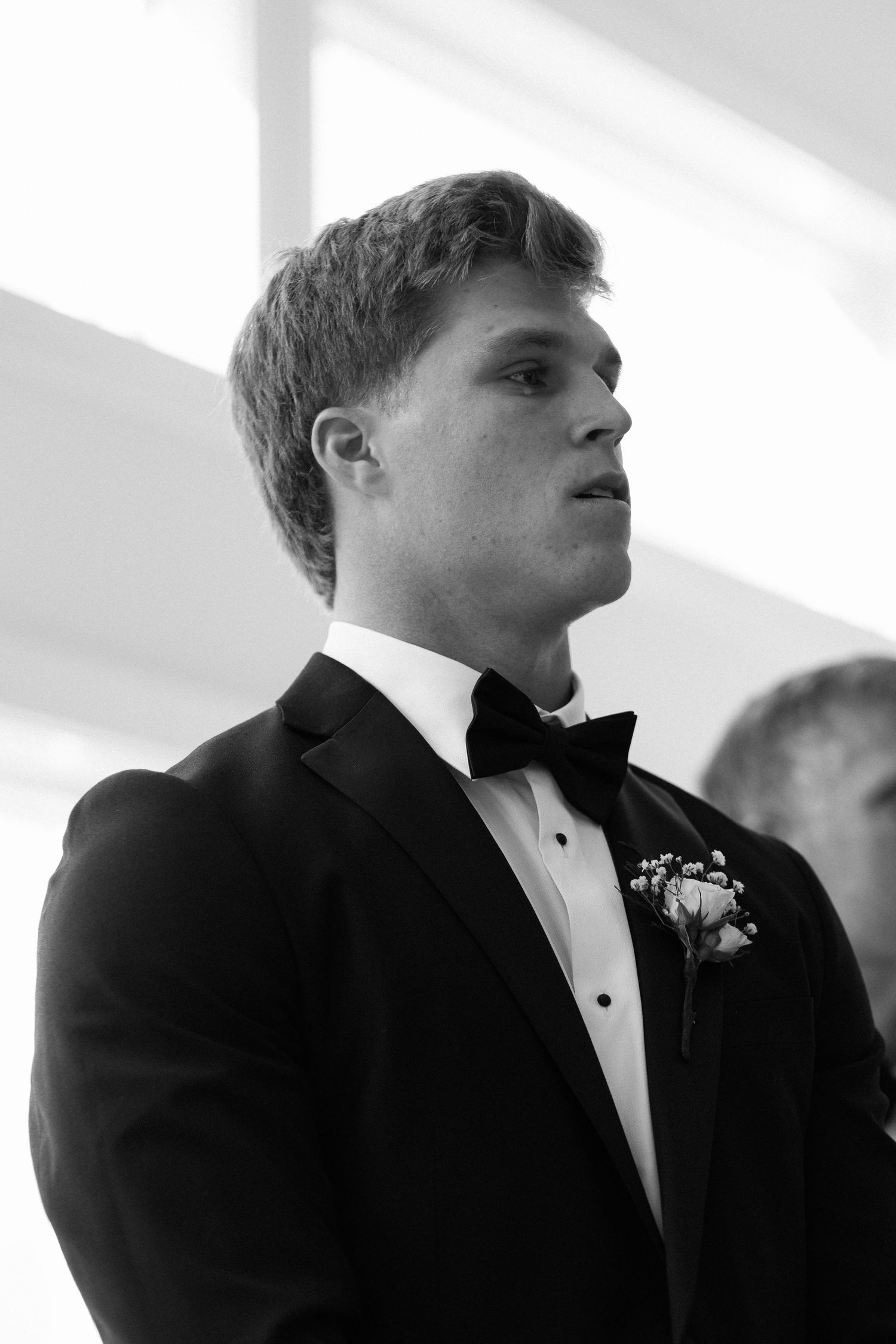 Black and white portrait of groom in black tuxedo with boutonniere during wedding ceremony.