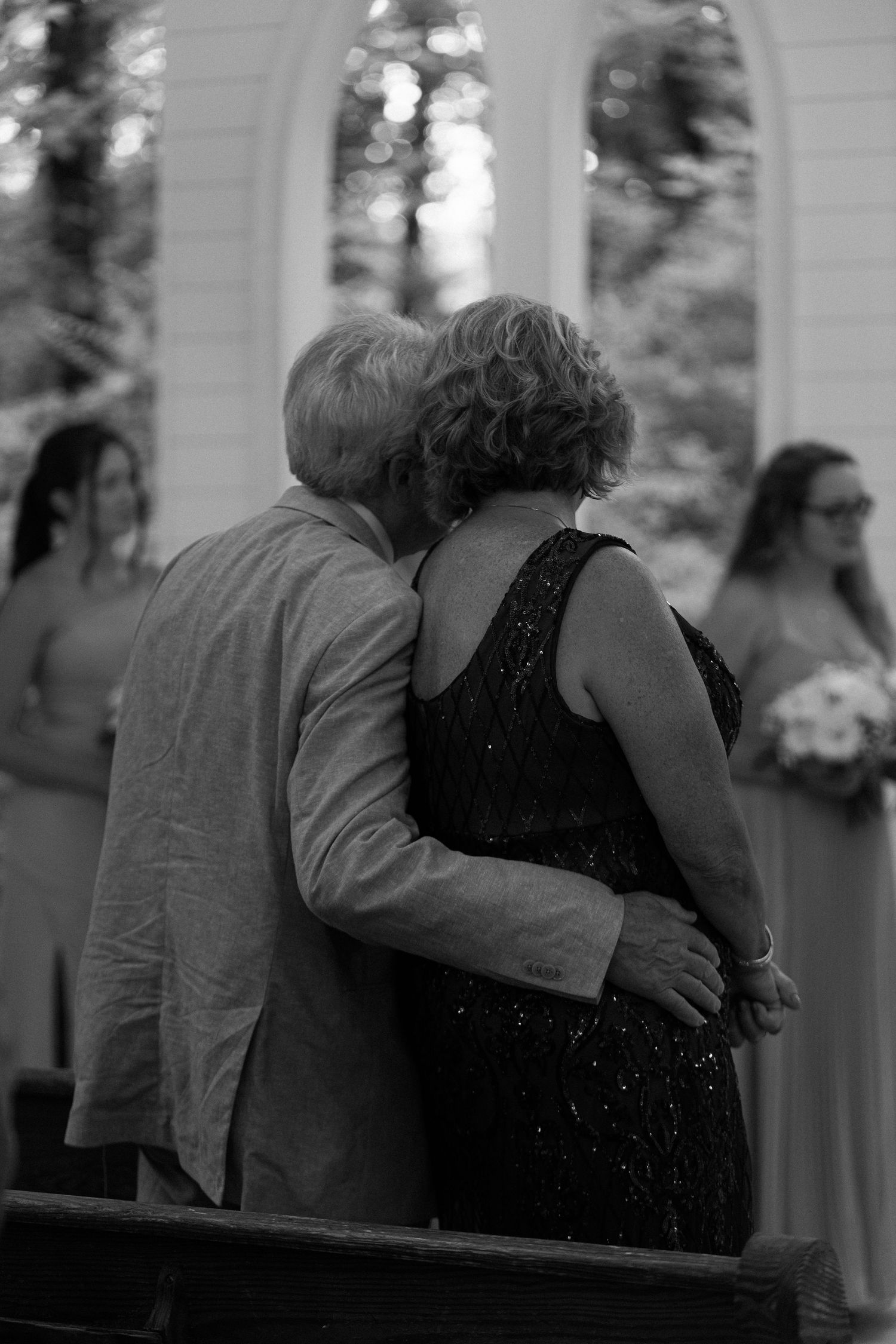 Emotional black and white photo of couple embracing at wedding ceremony with bridesmaids in background.