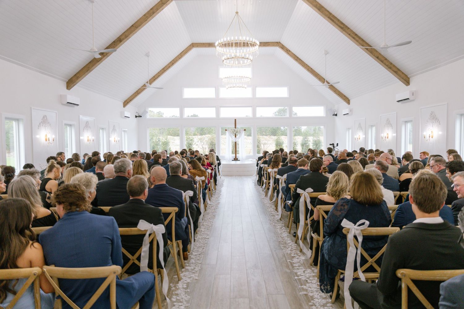 Guests fill wooden chairs during a wedding ceremony inside a bright white chapel with vaulted beam ceilings.