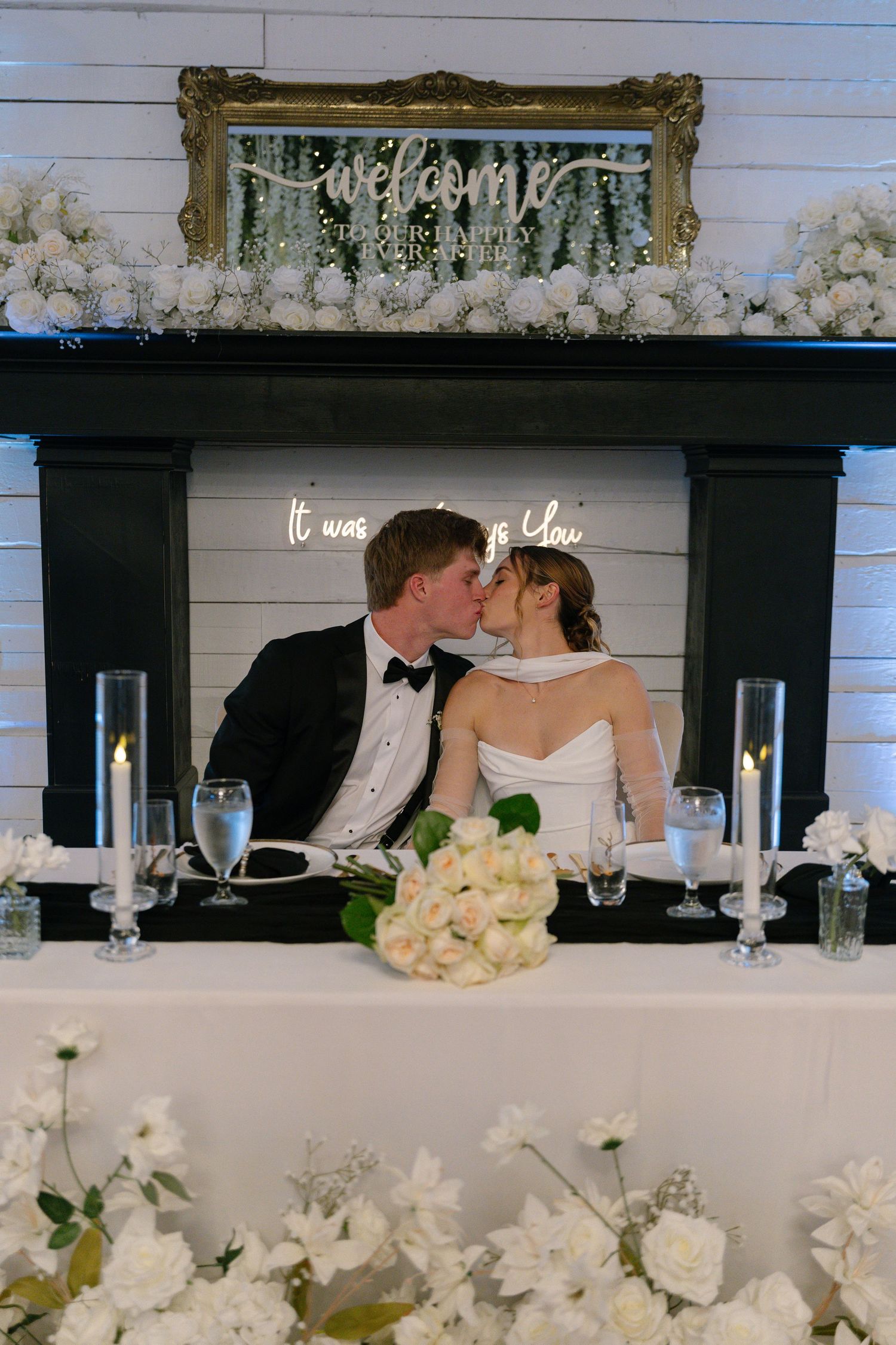 Bride and groom kiss at sweetheart table adorned with white roses and candles beside elegant fireplace.