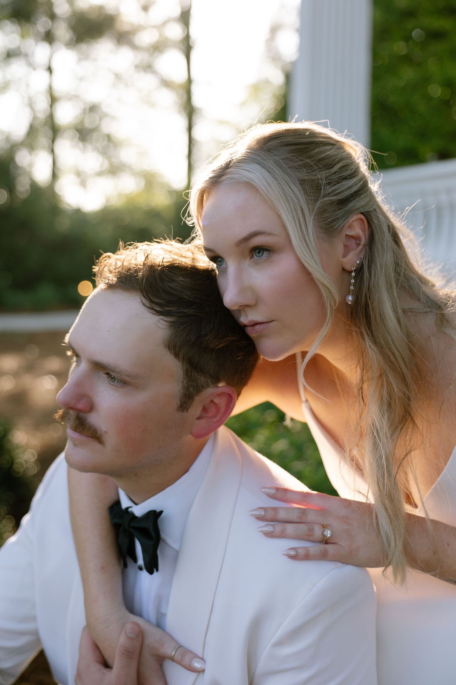 A bride in white leans over her groom in a white tuxedo outdoors, with soft golden sunlight and greenery behind them.