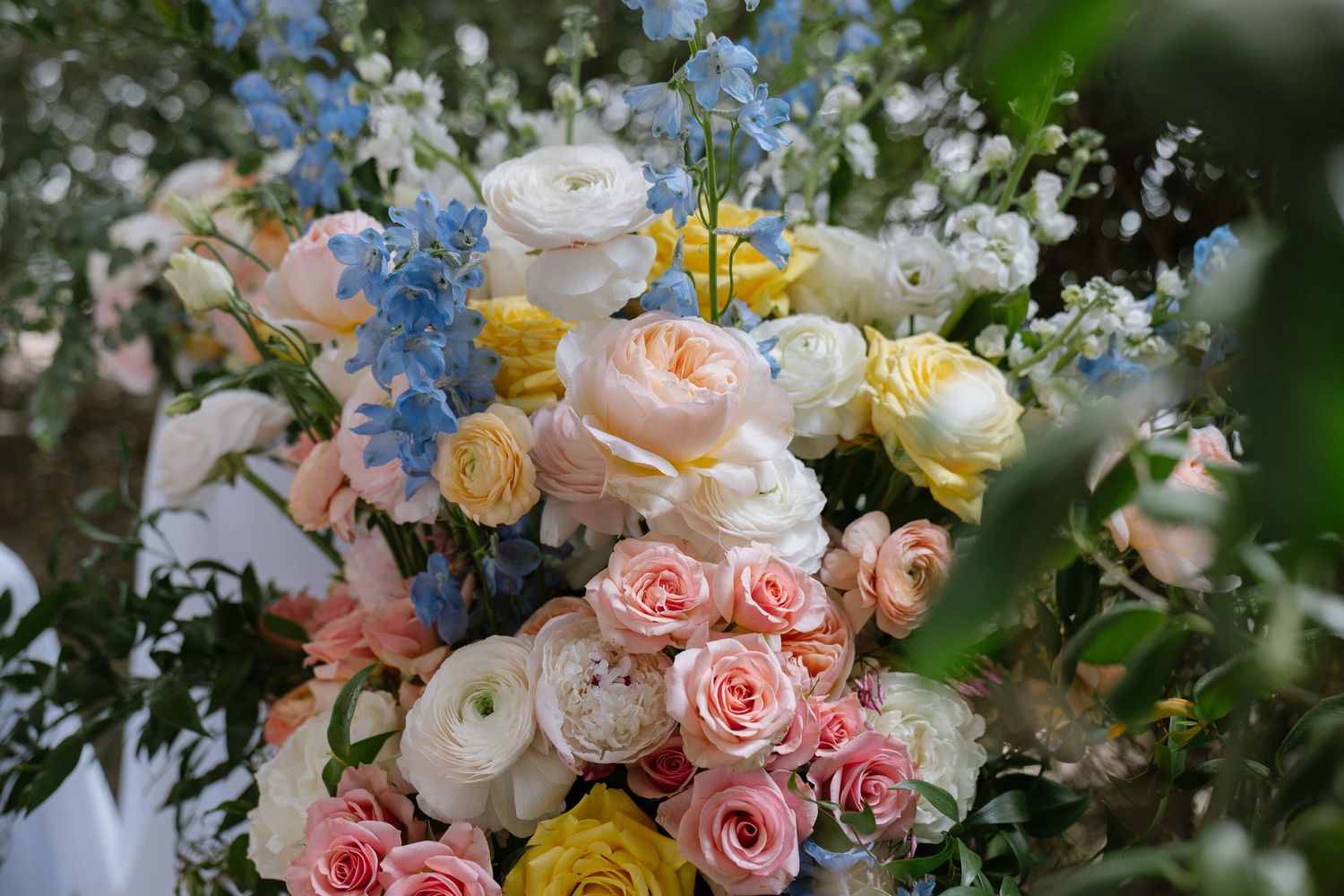 Lush wedding bouquet featuring pink, white, yellow roses, blue delphiniums, and ranunculus arranged in a romantic garden style.