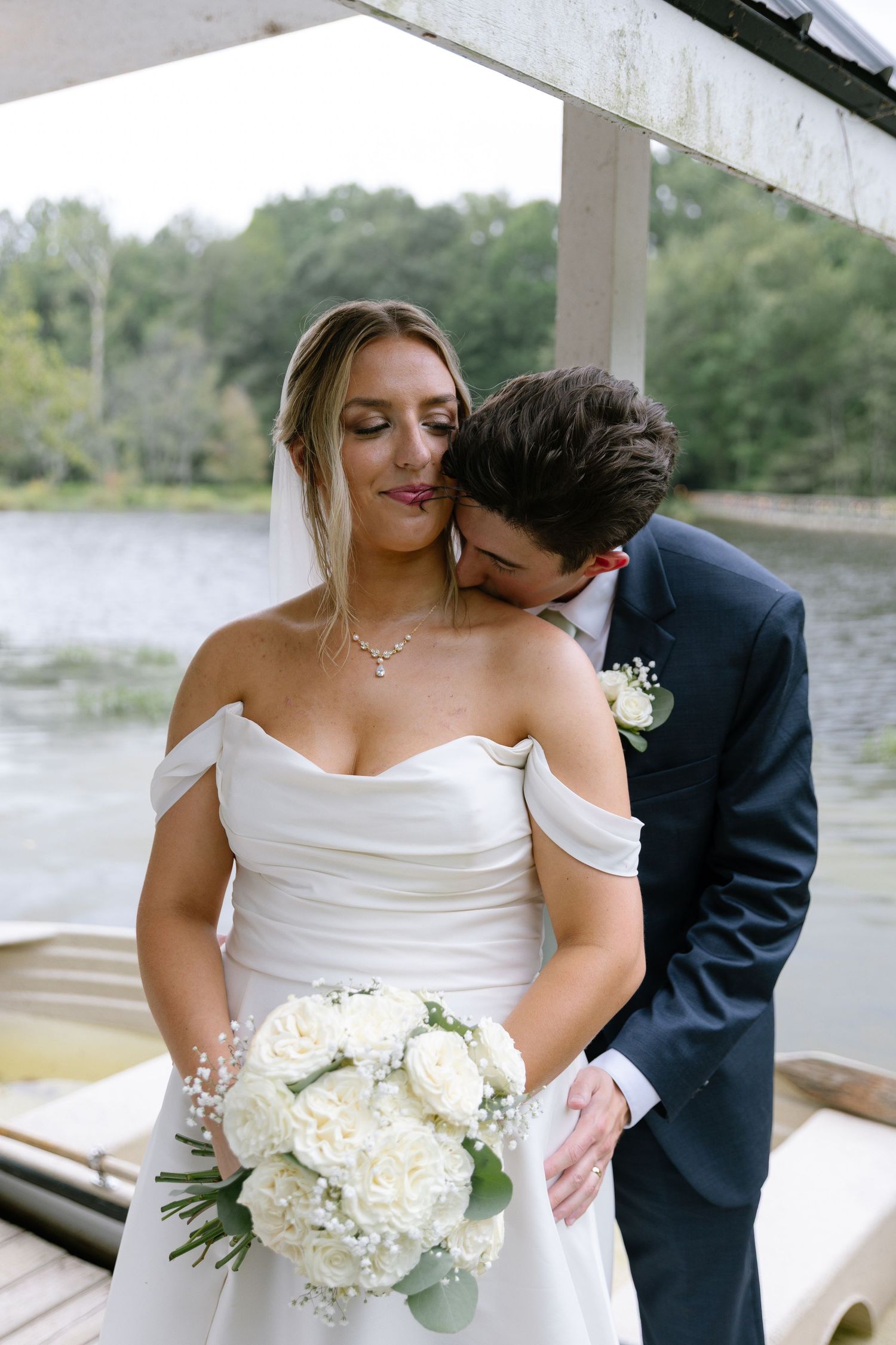 Groom in navy suit kissing bride on neck as she smiles holding white rose bouquet at lakeside boat dock.