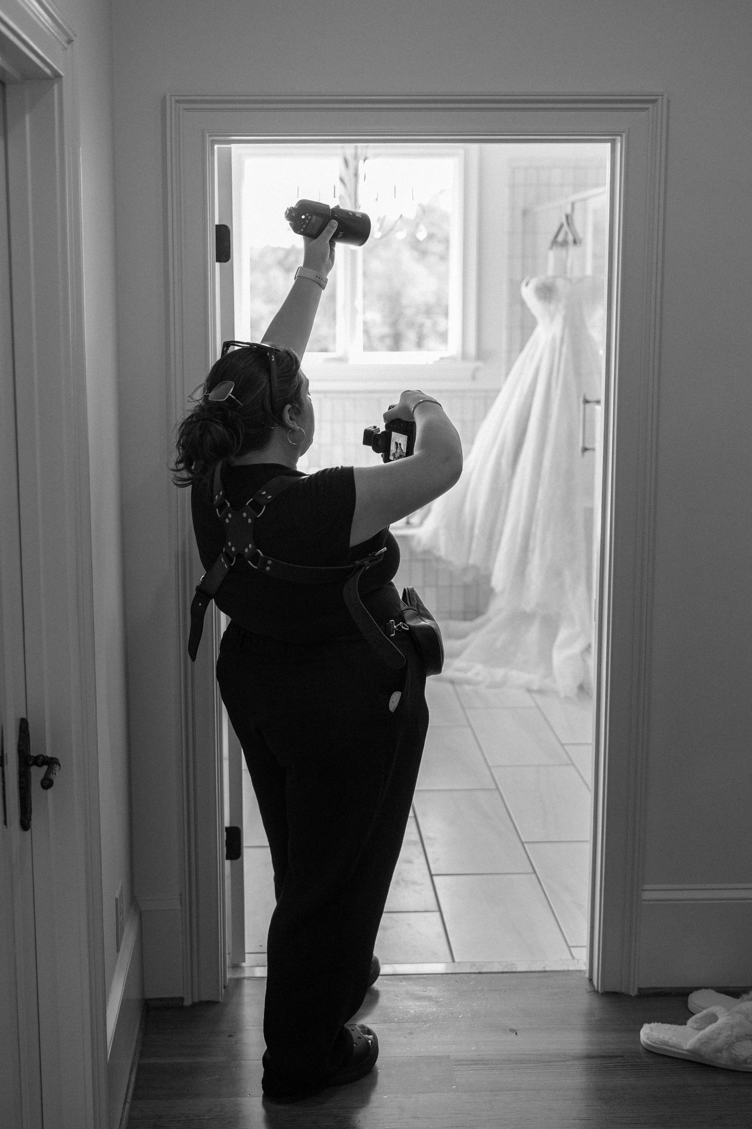 Black and white photo of a female photographer holding cameras in a doorway with a wedding dress hanging in the background.