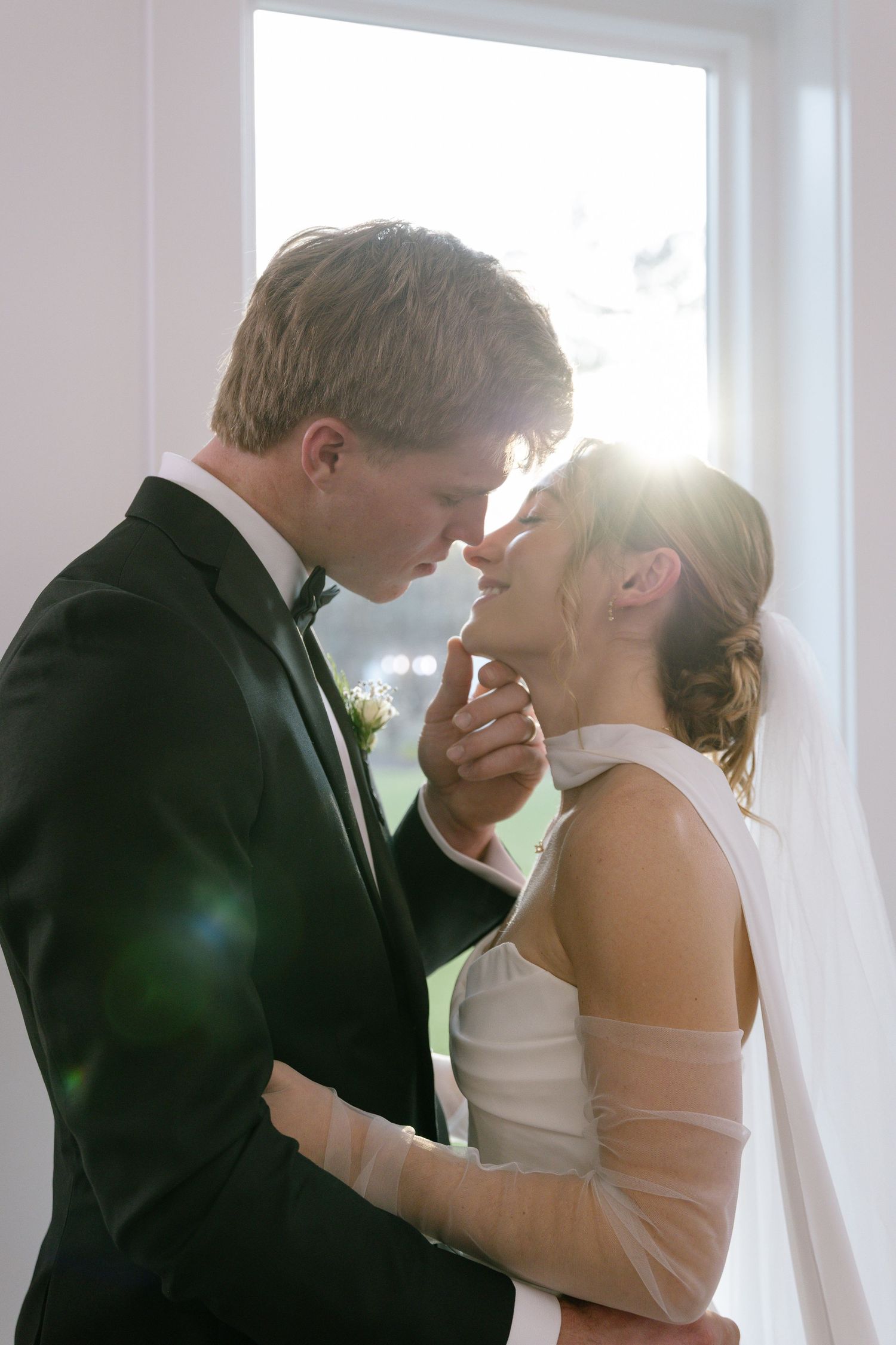 Bride and groom share an intimate moment near a sunlit window, groom gently holding bride's face.