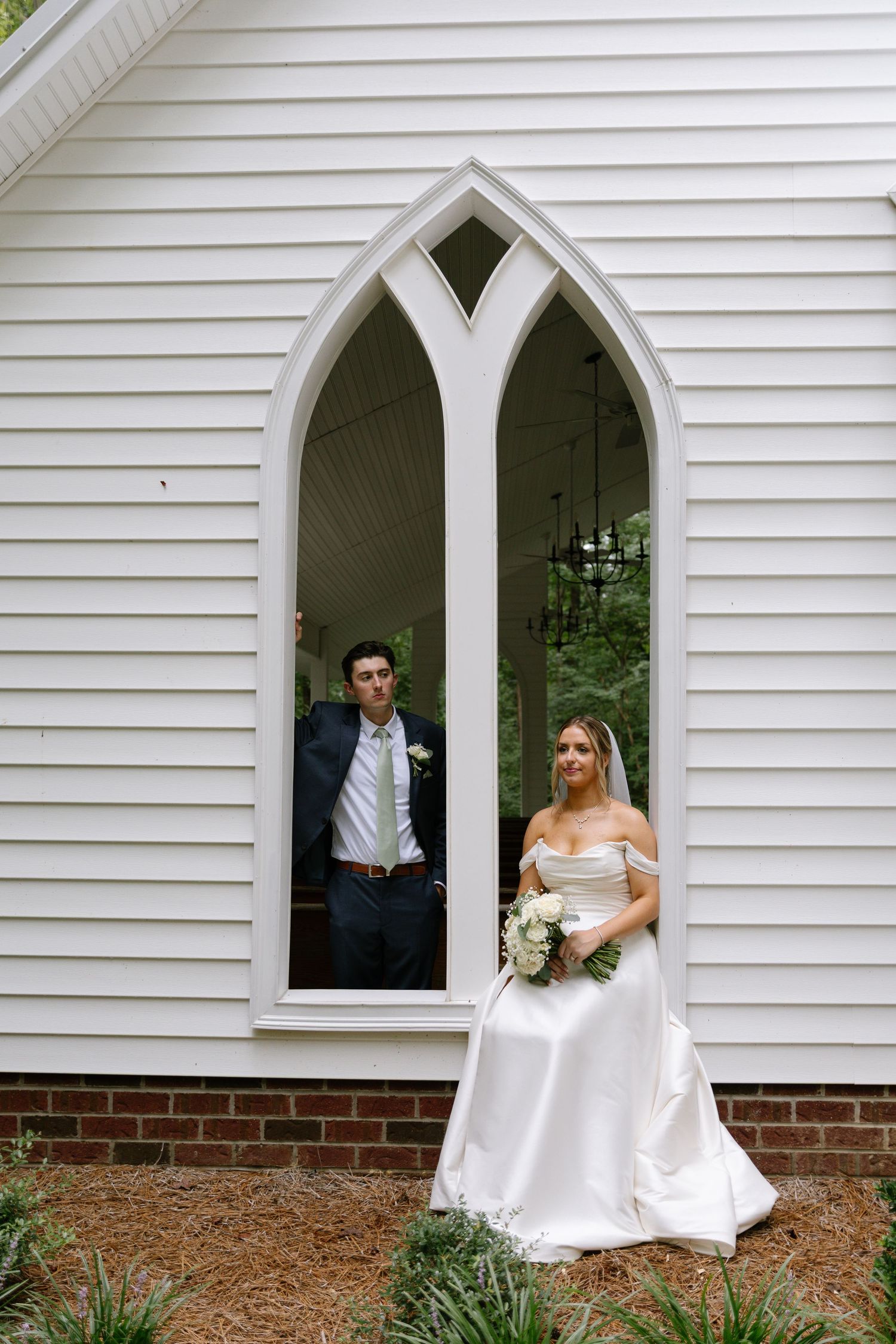 Bride holding white bouquet poses by gothic arched window while groom stands inside a white chapel.