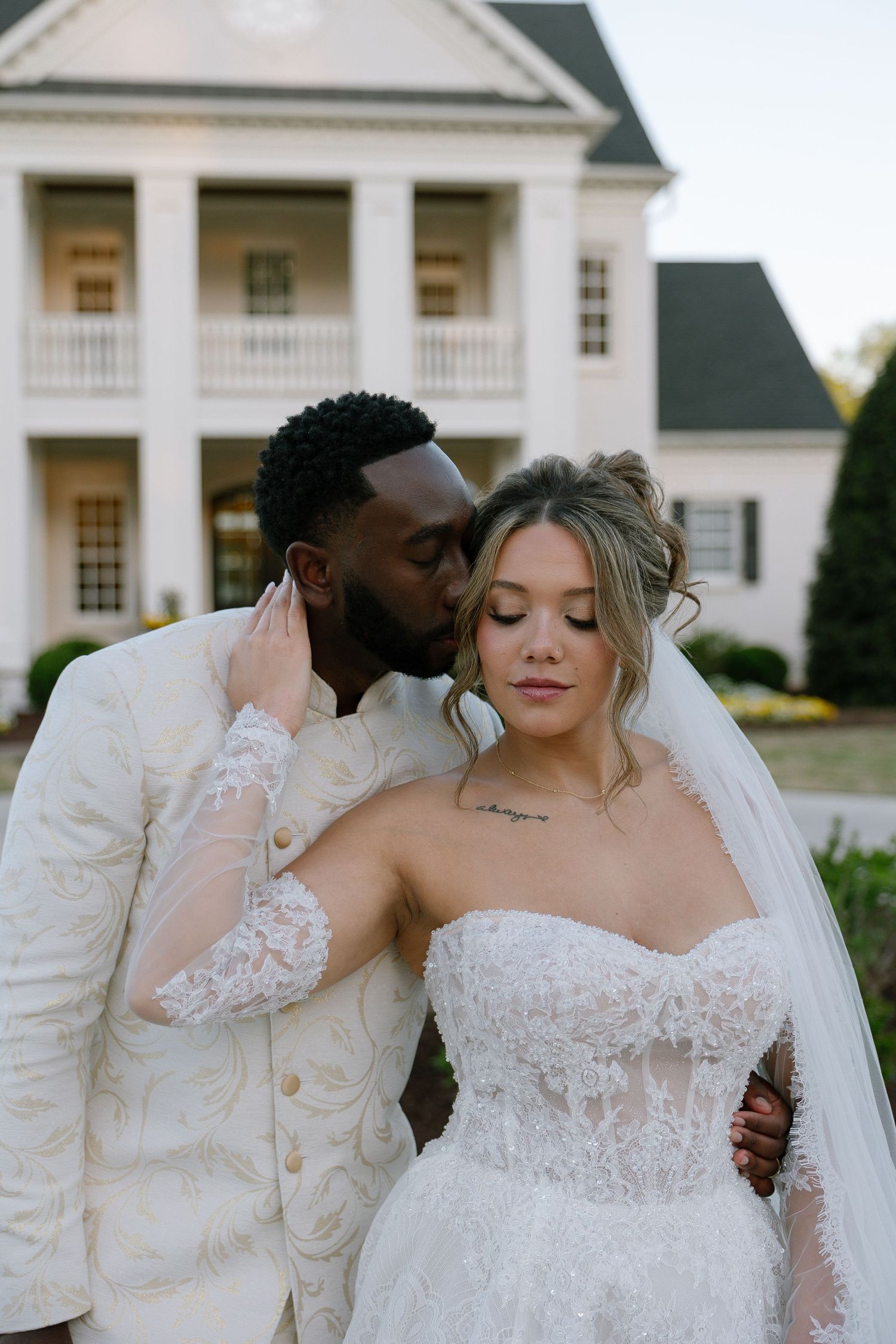 Groom in ivory suit kissing bride in strapless lace wedding gown outside elegant white mansion.