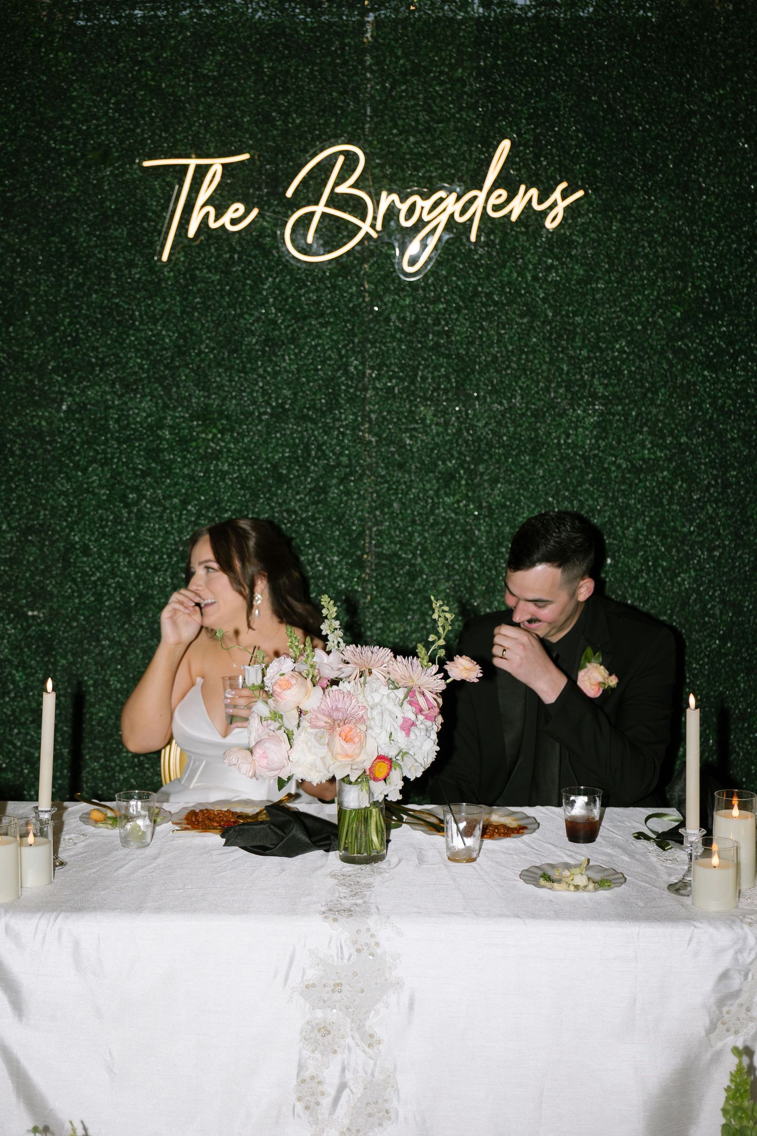 Bride and groom sit at sweetheart table beneath glowing The Brogdens neon sign on lush green hedge wall.