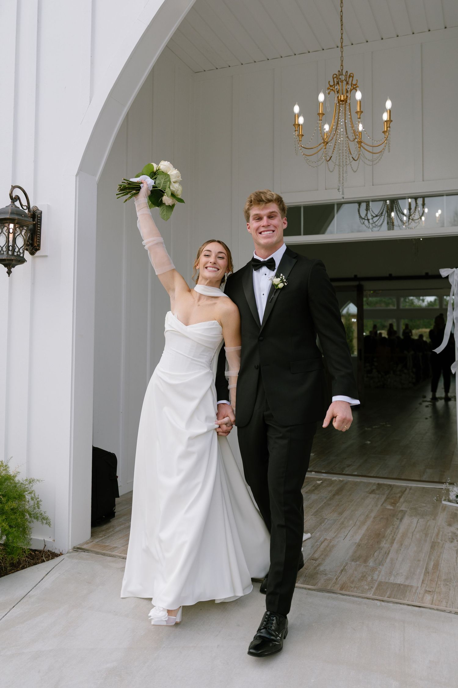 Joyful newlyweds exit white chapel arch, bride raising bouquet in celebration with groom by her side.