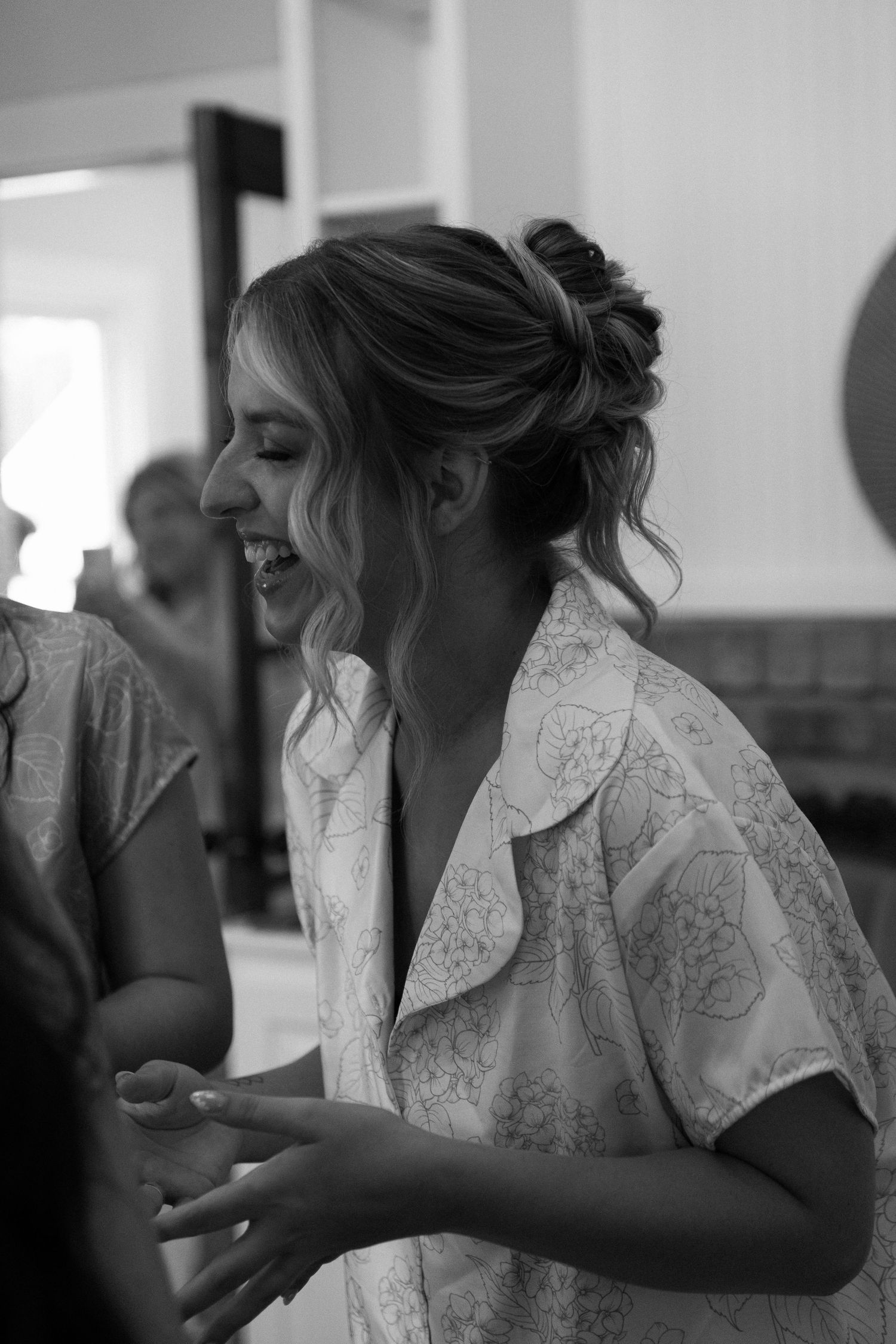 Black and white photo of a laughing bride in a floral robe with an elegant updo hairstyle during wedding preparations.