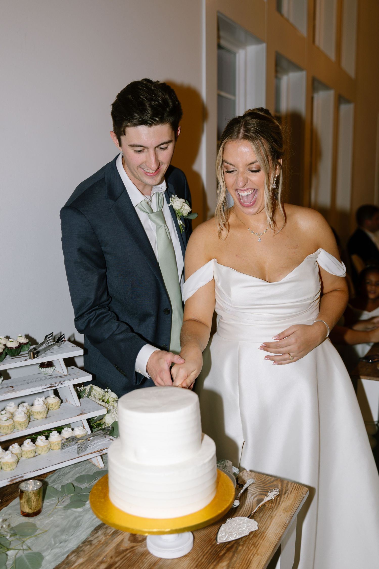 Laughing bride and groom cutting two-tier white wedding cake together at elegant indoor wedding reception.