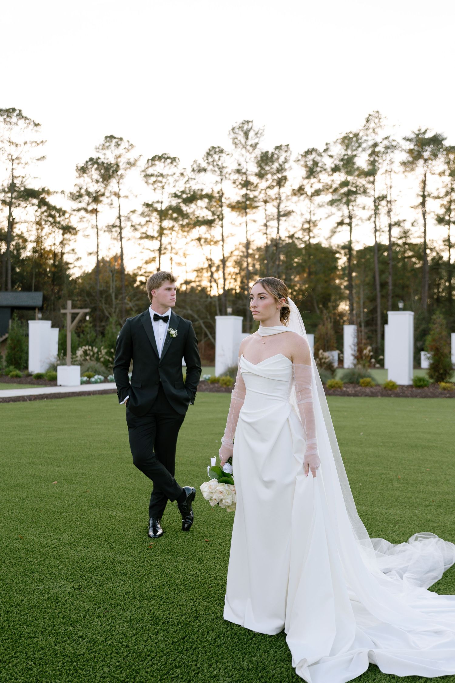 Bride in elegant white halter gown with cathedral train poses with groom in black tuxedo outdoors.
