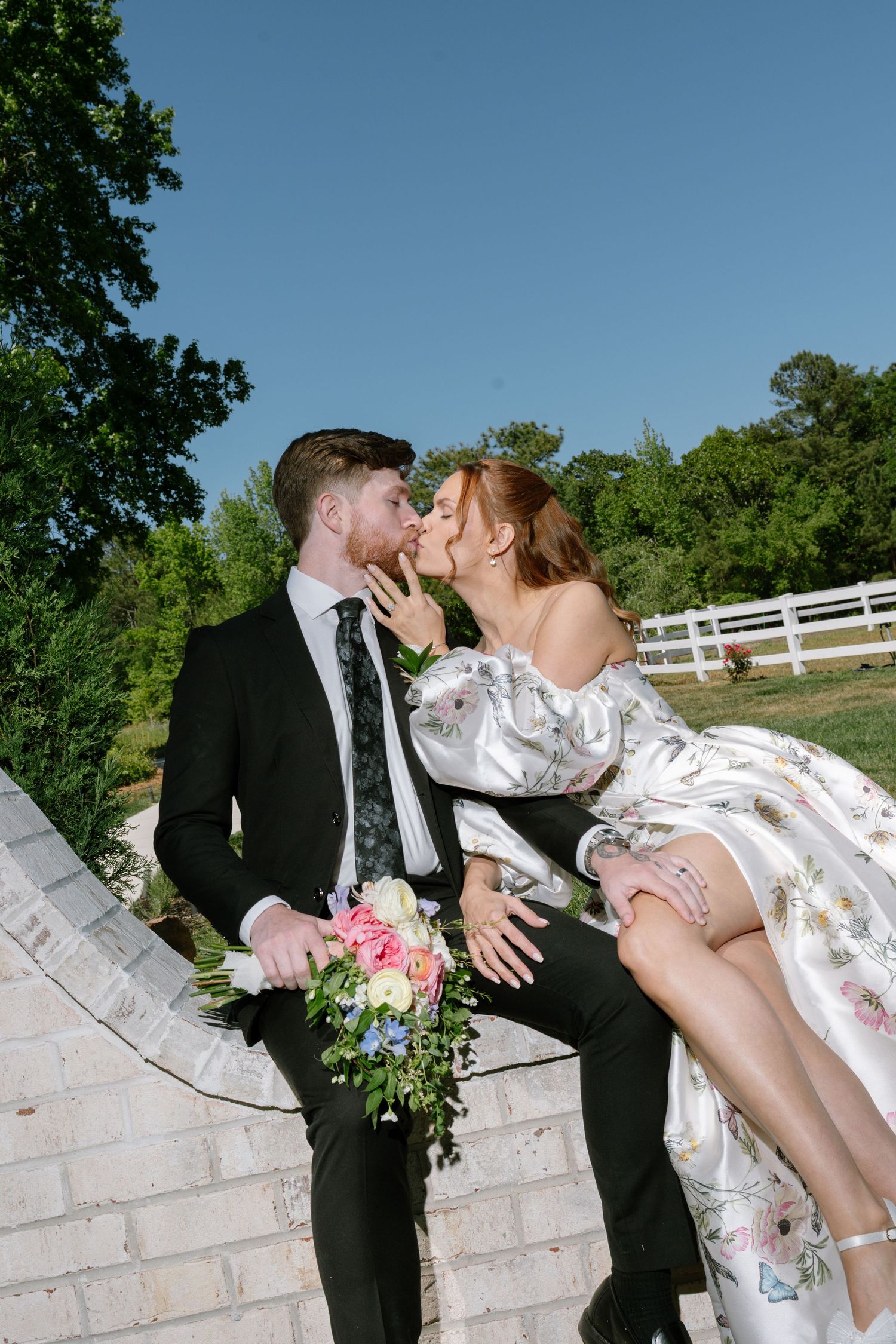Bride and groom share a romantic kiss outdoors, with the bride in a floral gown holding a bouquet on a sunny day.