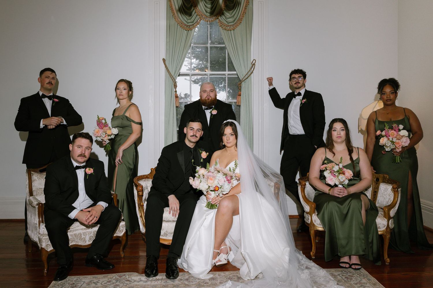Wedding party poses elegantly in a historic venue, bride and groom centered with bridesmaids in green gowns and groomsmen in tuxedos.
