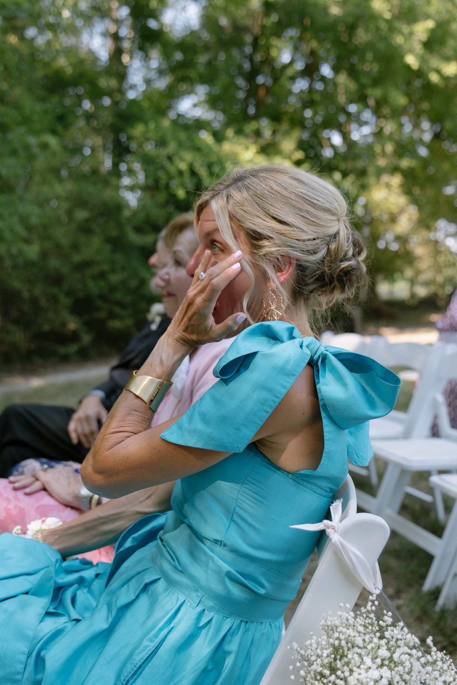Woman in turquoise bow dress wiping tears at outdoor wedding ceremony, seated among guests.