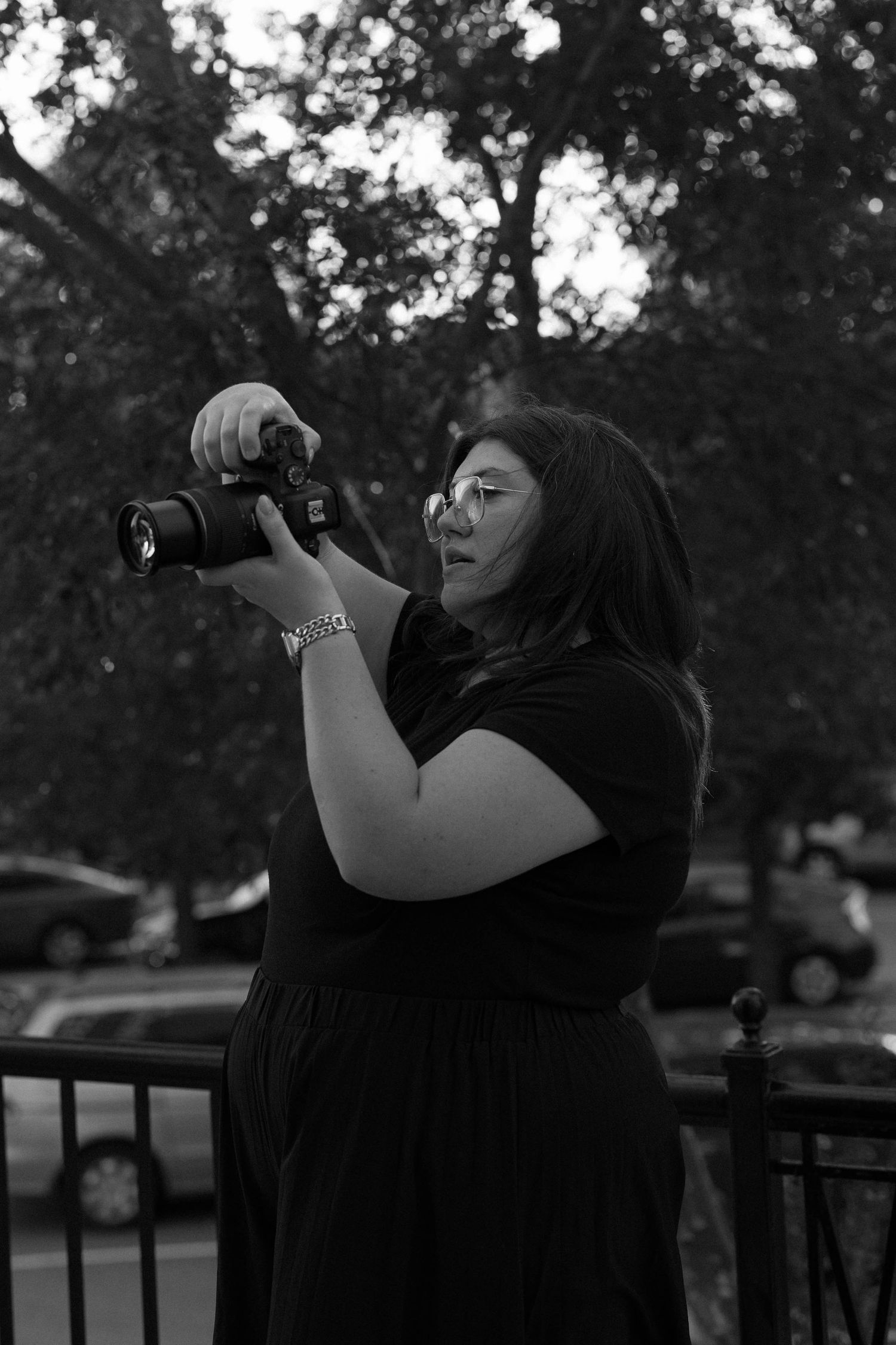 Black and white photo of a woman holding a DSLR camera outdoors, wearing glasses and a dark dress.