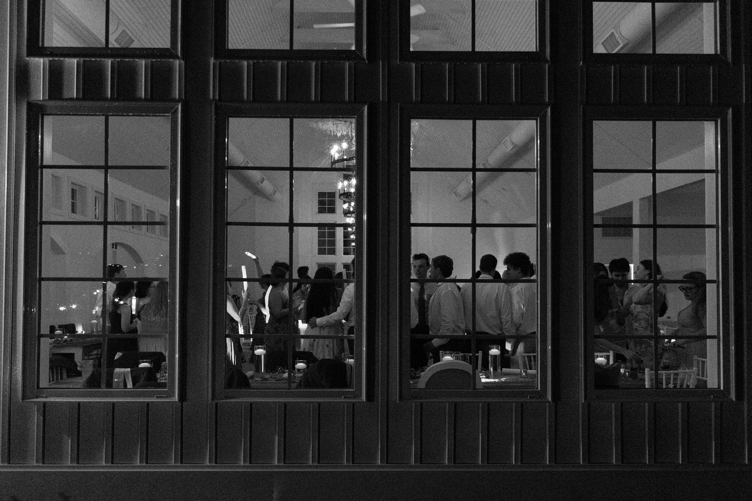 Black and white photo of wedding reception guests gathered inside venue viewed through large windows.