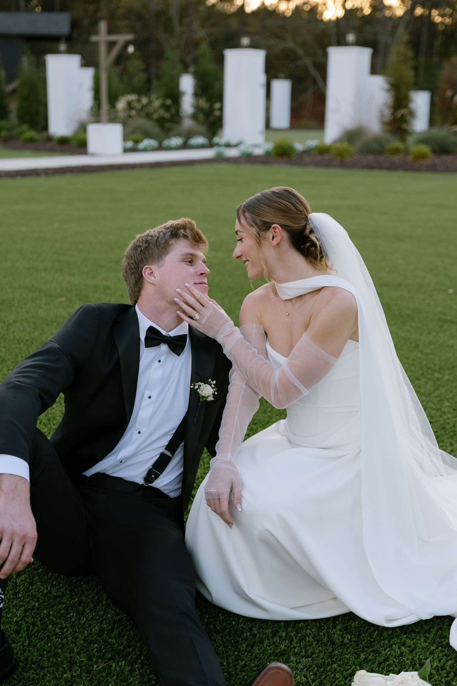 Bride and groom sitting on green lawn at sunset, sharing tender moment at outdoor wedding venue.