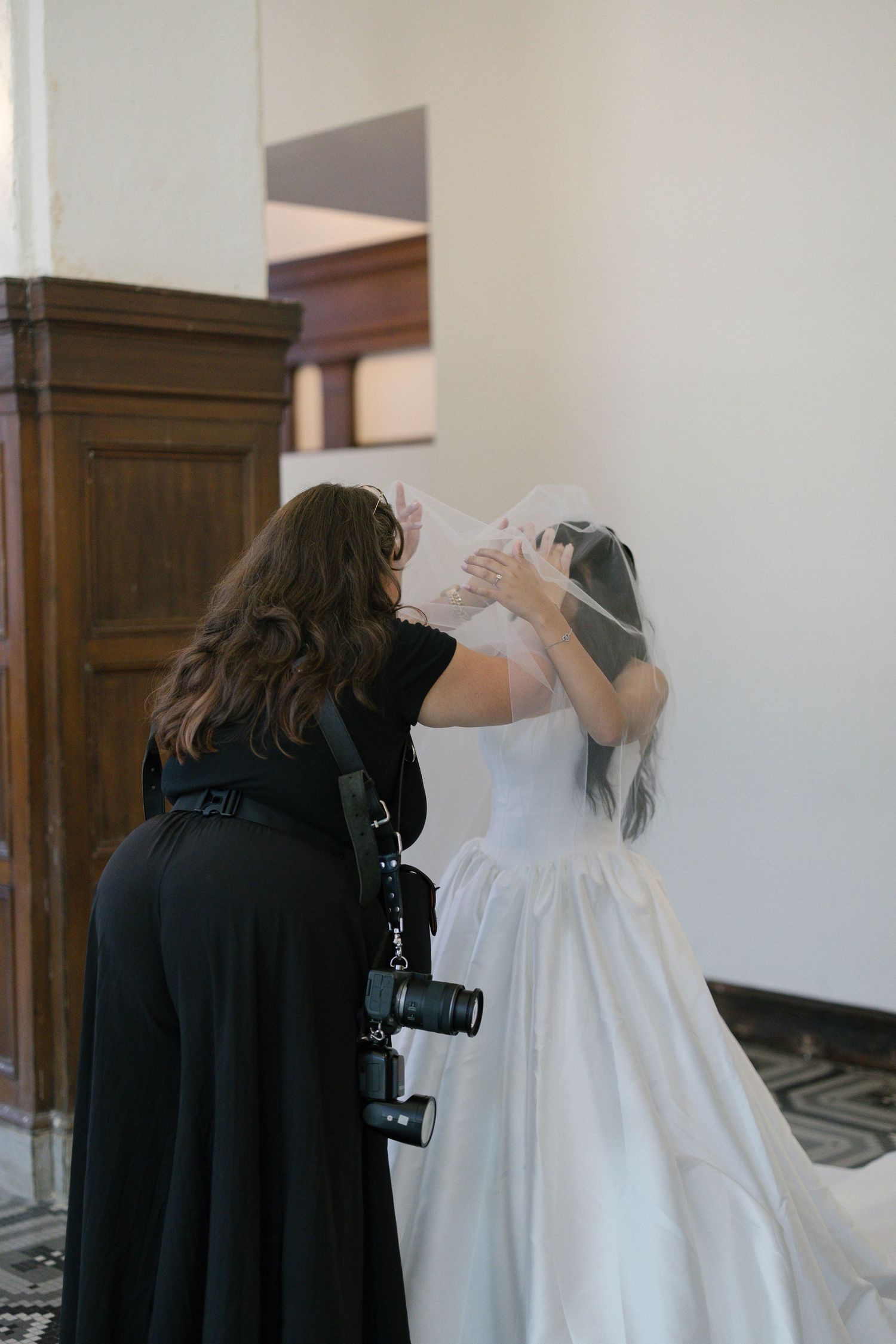 Wedding photographer in black adjusting bride's veil before ceremony in elegant venue.