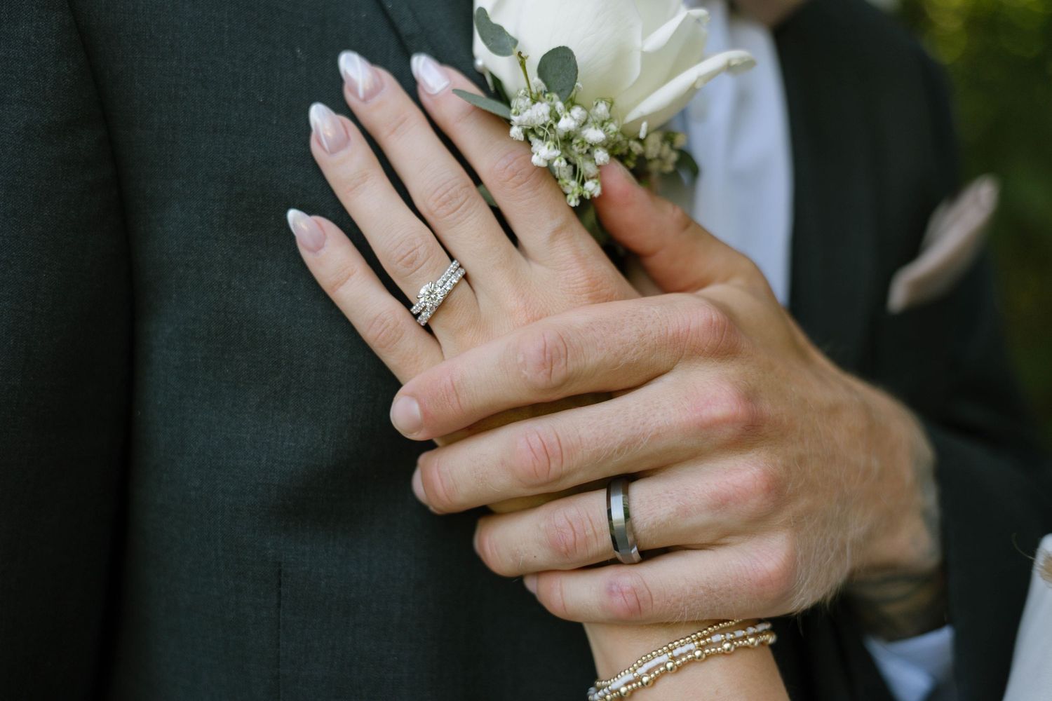 Newlyweds displaying wedding rings, bride with French manicure and bracelet, groom in dark suit with white boutonniere.