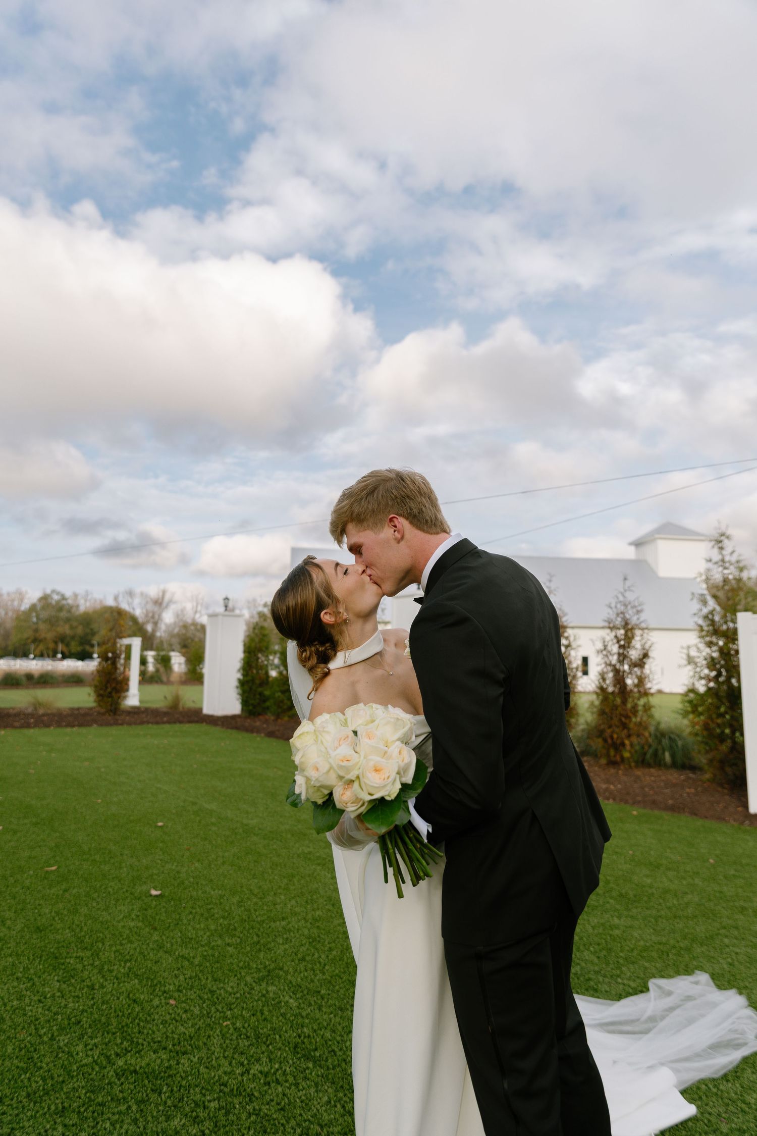 Bride and groom sharing a romantic kiss on green lawn with white barn venue and cloudy sky.