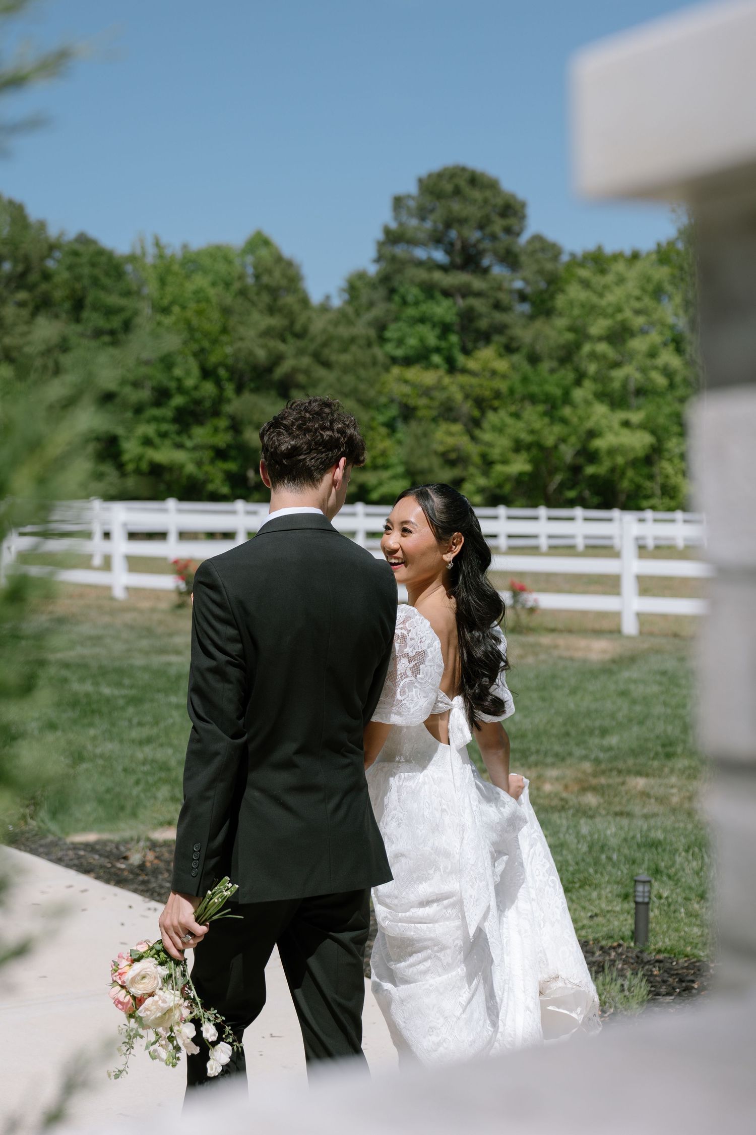 Bride in lace gown and groom in black tuxedo walk hand-in-hand outdoors near white fence on sunny wedding day.