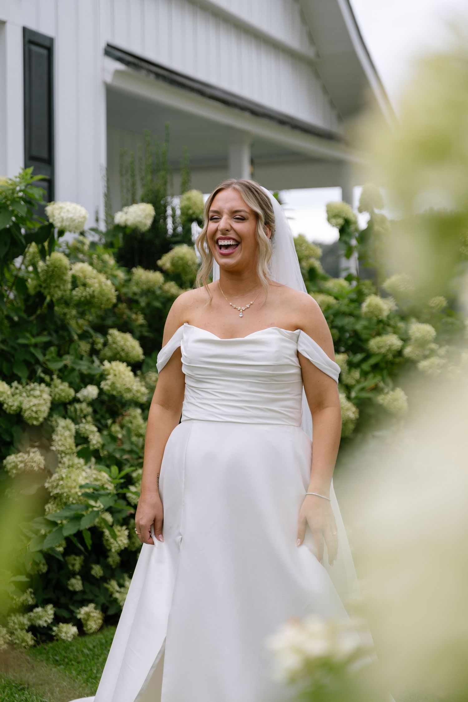 Laughing bride in off-shoulder white gown stands before lush hydrangeas outside a white farmhouse.
