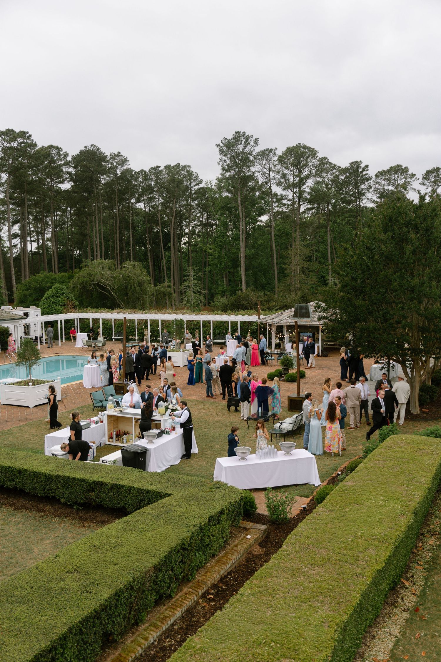 Elegant outdoor wedding reception with guests mingling on manicured grounds near a pool, surrounded by tall pine trees.