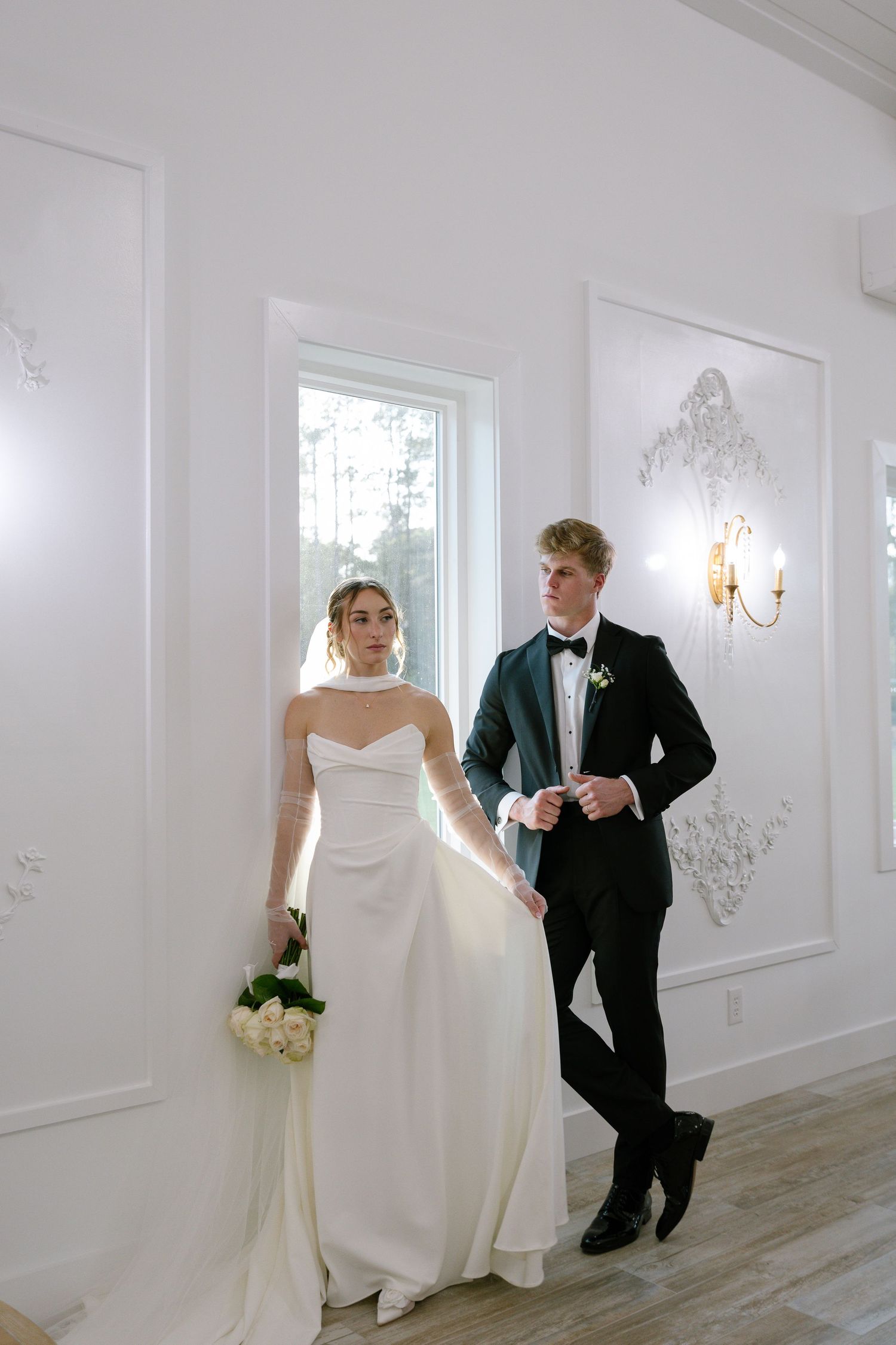 Bride in white strapless gown and groom in black tuxedo pose together in elegant white venue interior.
