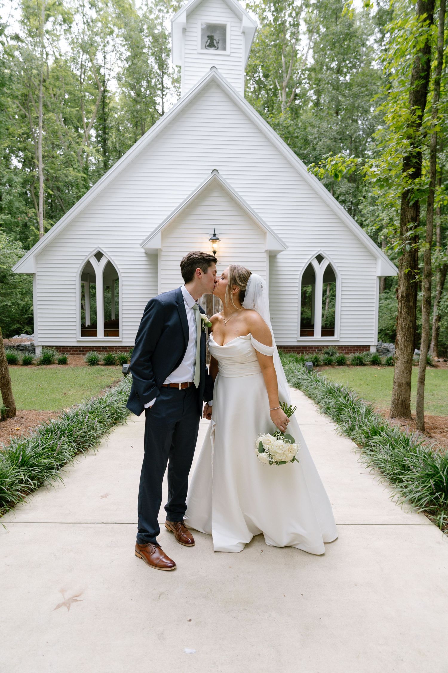 Bride and groom share a romantic kiss on the pathway in front of a charming white chapel surrounded by trees.