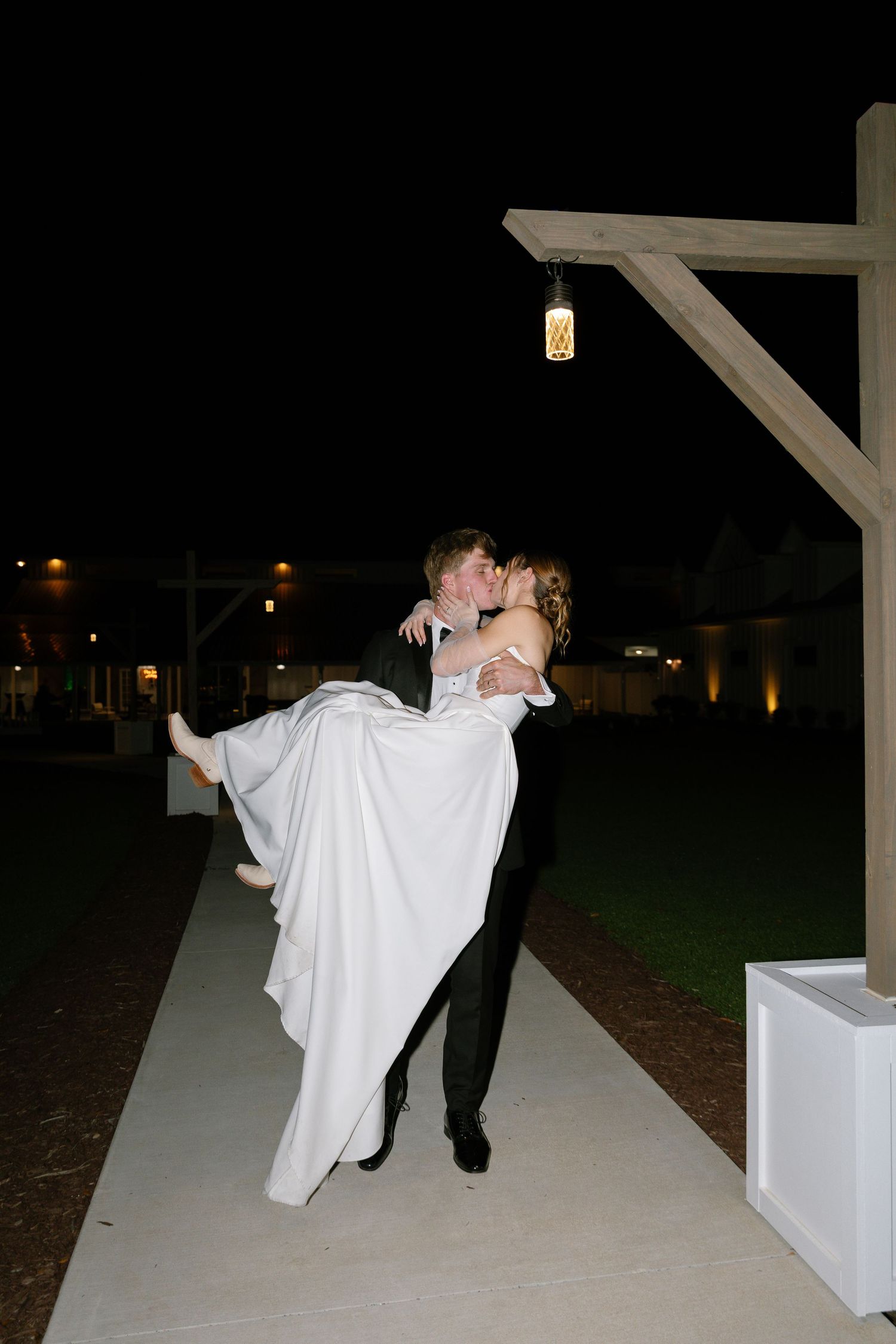 Groom lifts and kisses bride in white wedding gown outdoors at night under warm string lights.