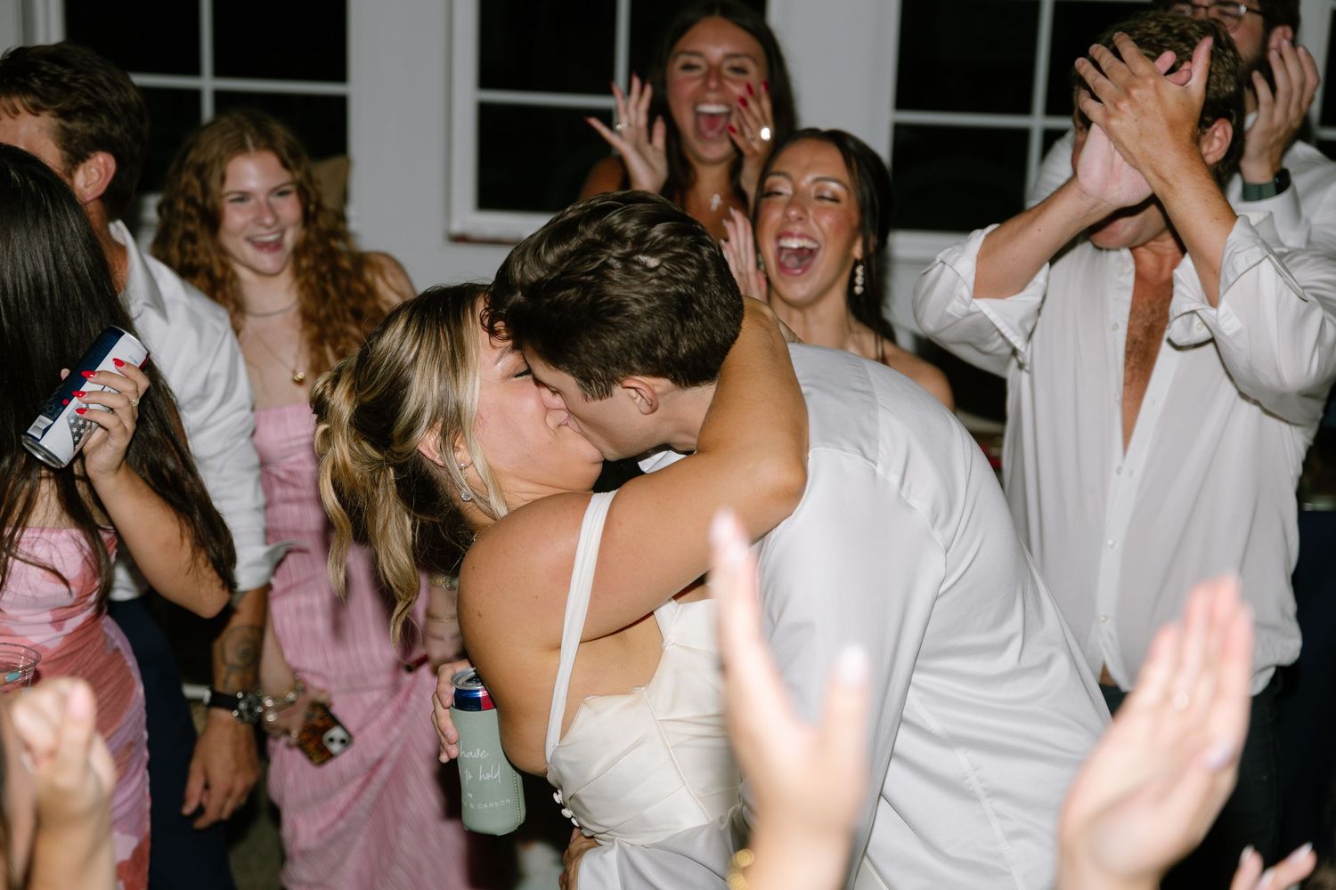 Bride and groom share passionate kiss on dance floor surrounded by cheering wedding reception guests.