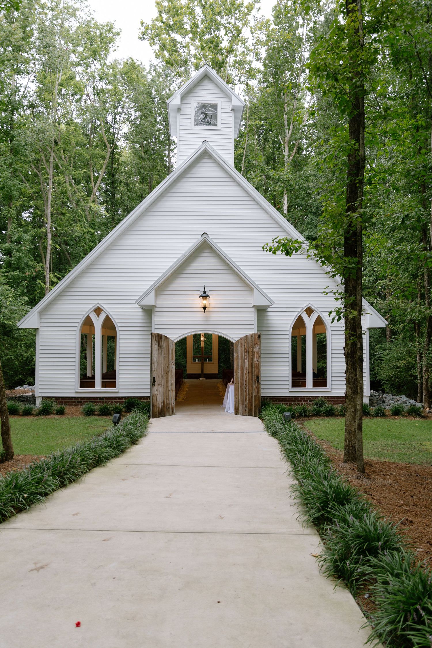 Elegant white chapel wedding venue with Gothic arch windows and wooden doors surrounded by tall trees.