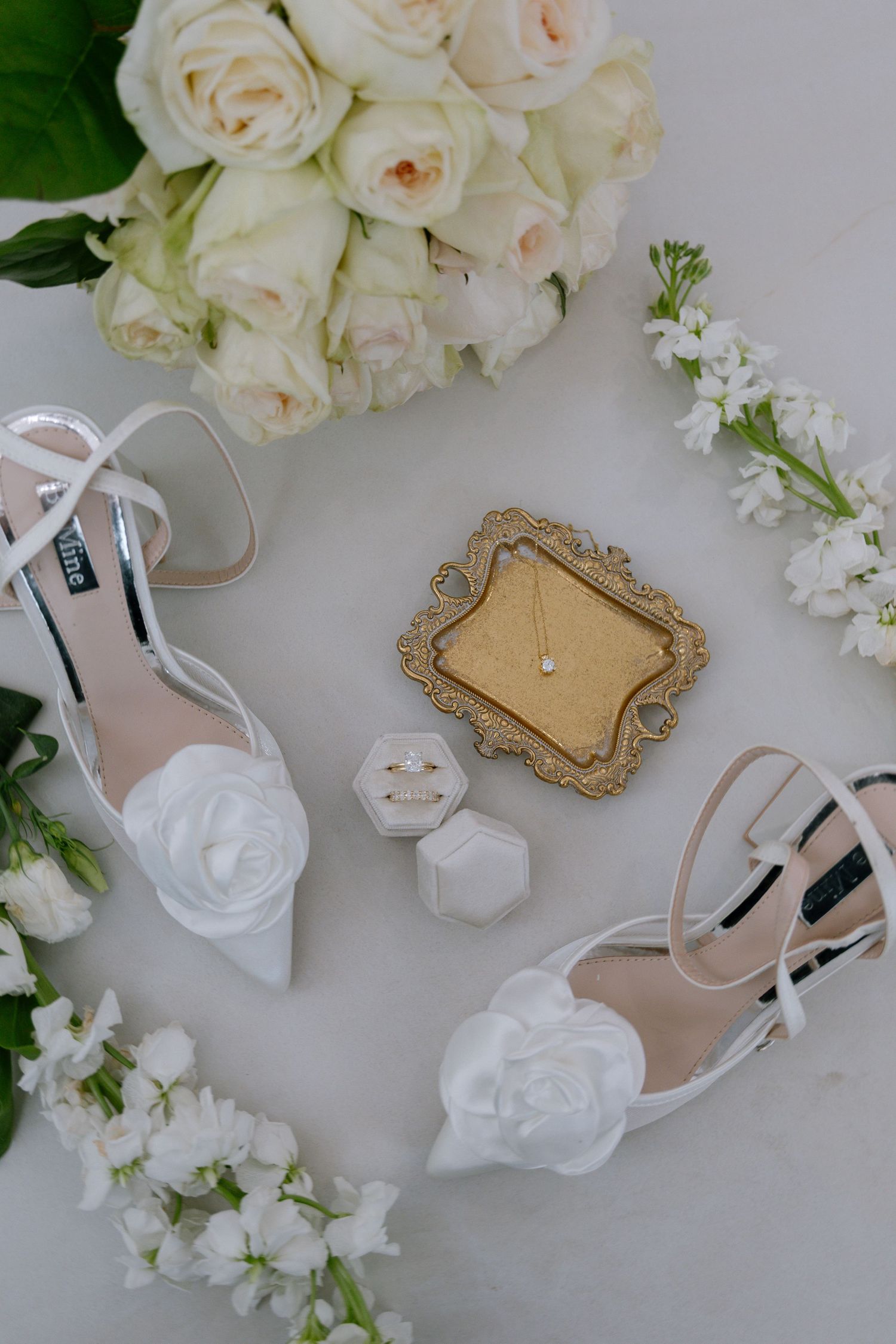 White bridal heels with floral detail, ring box, gold jewelry tray, and white roses arranged on marble surface.
