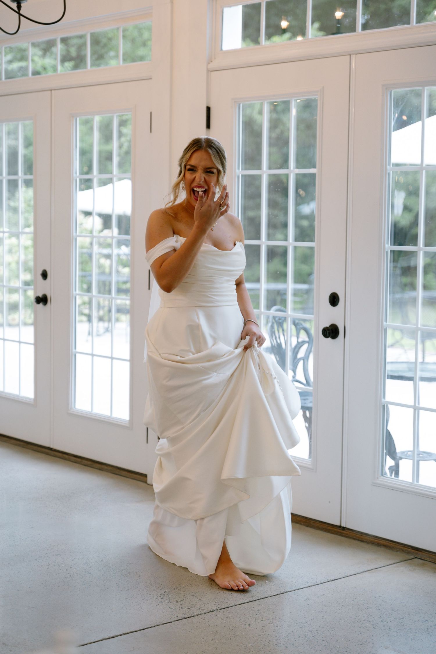 Happy barefoot bride in a white off-shoulder gown laughing near elegant French doors.