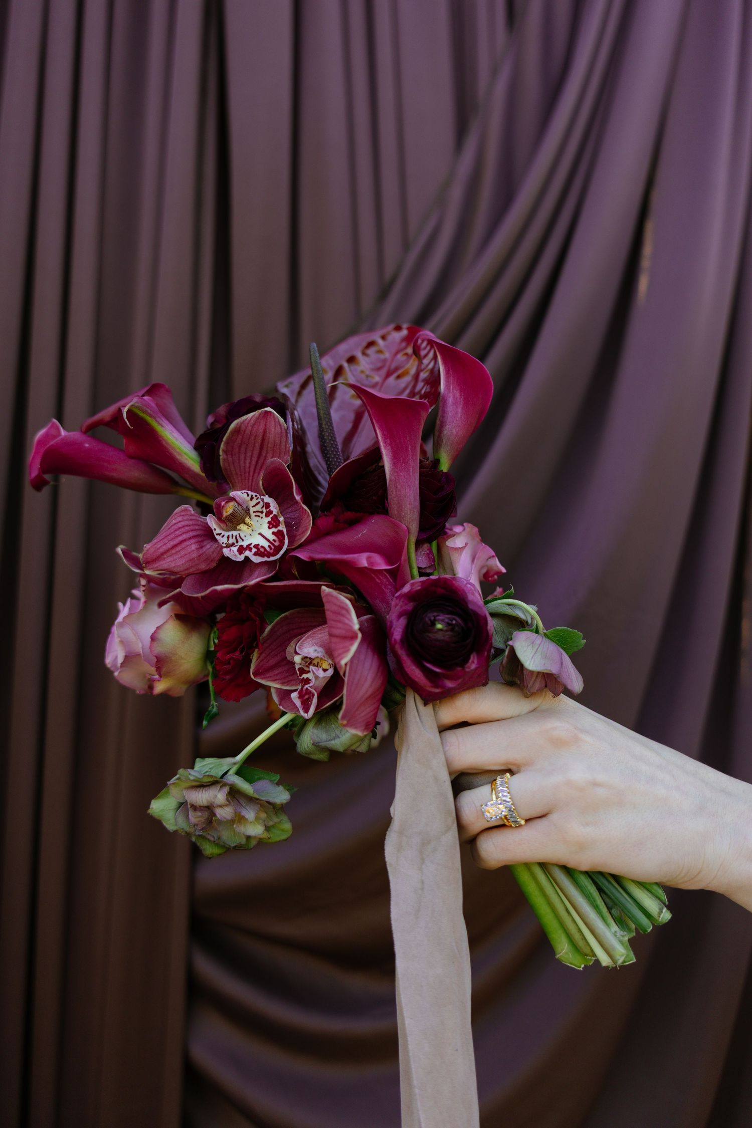 Hand holding a deep burgundy bridal bouquet with orchids and calla lilies against a draped purple backdrop.