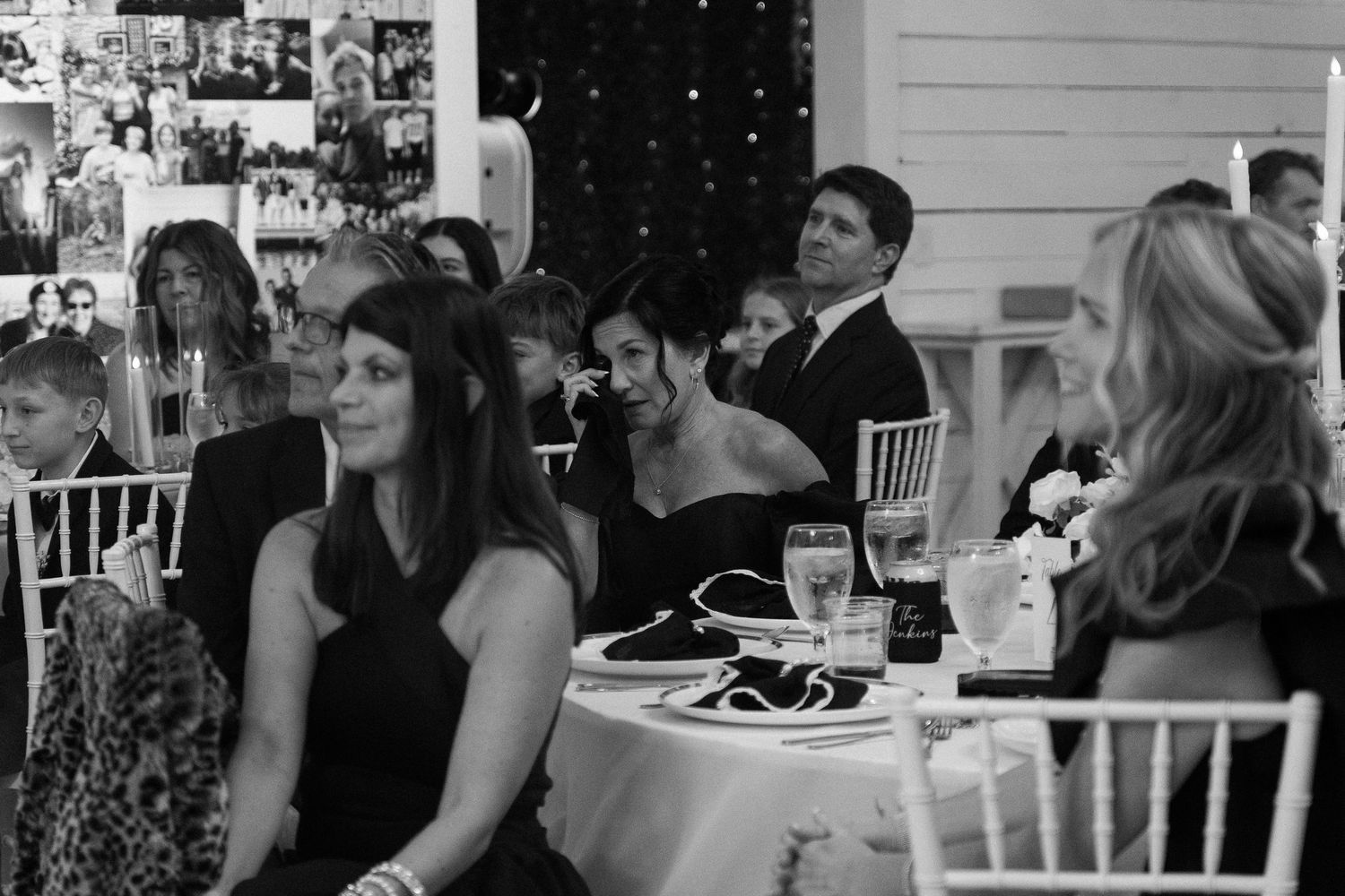Wedding guests seated at round tables during reception in black and white photo with photo wall backdrop.