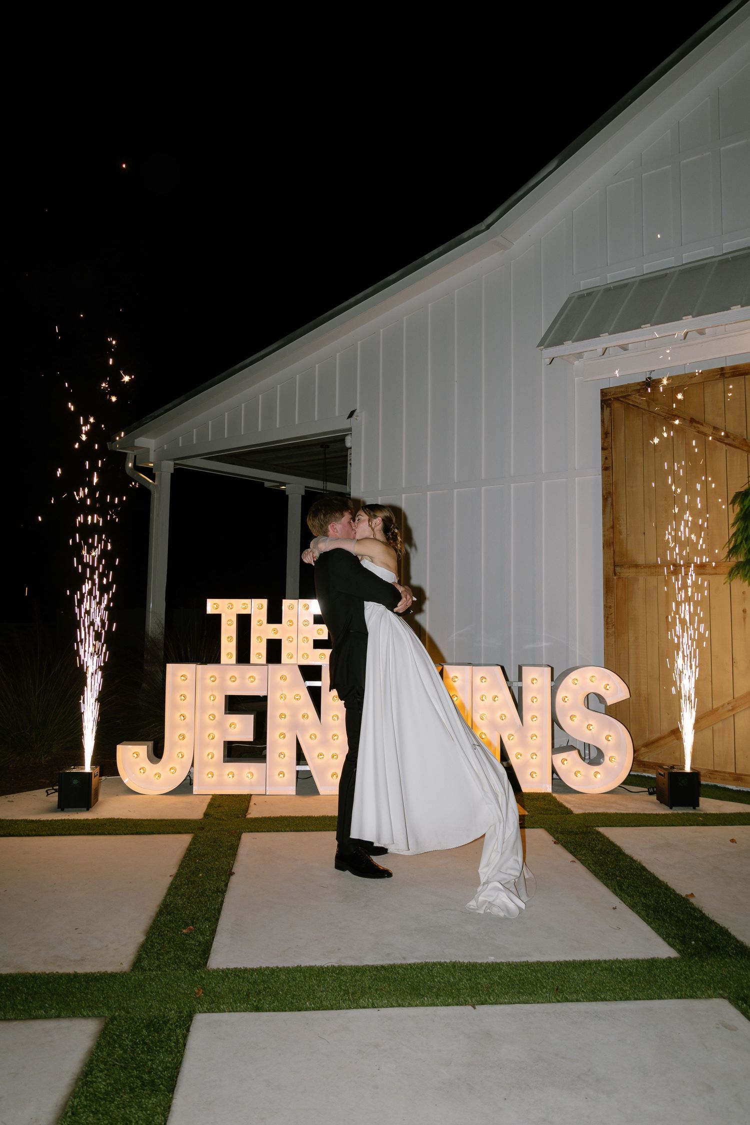 Newlyweds kiss in front of illuminated marquee letters spelling THE JENKINS with cold sparklers at night.