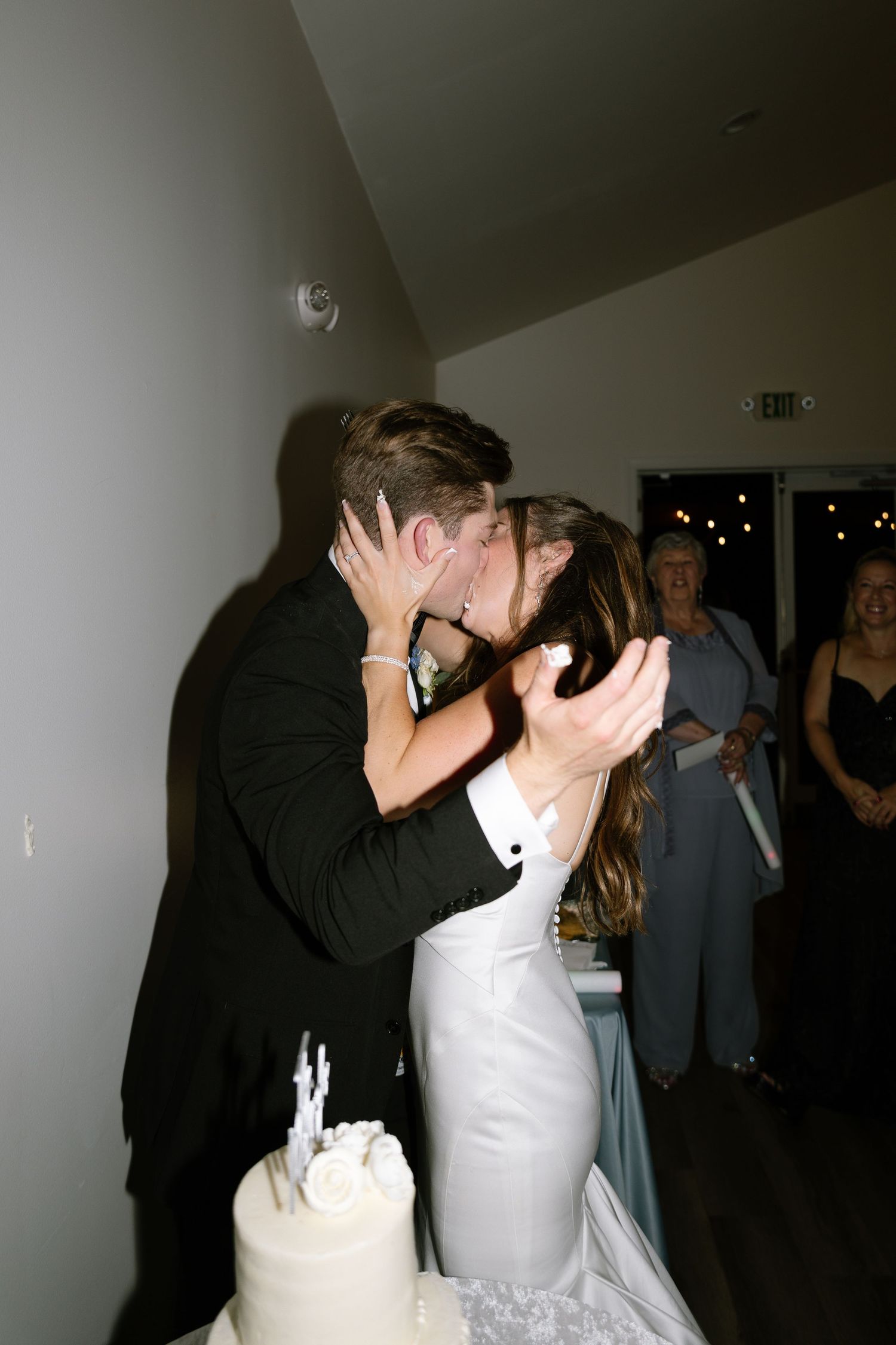Bride and groom share a romantic kiss beside their white wedding cake during their reception.