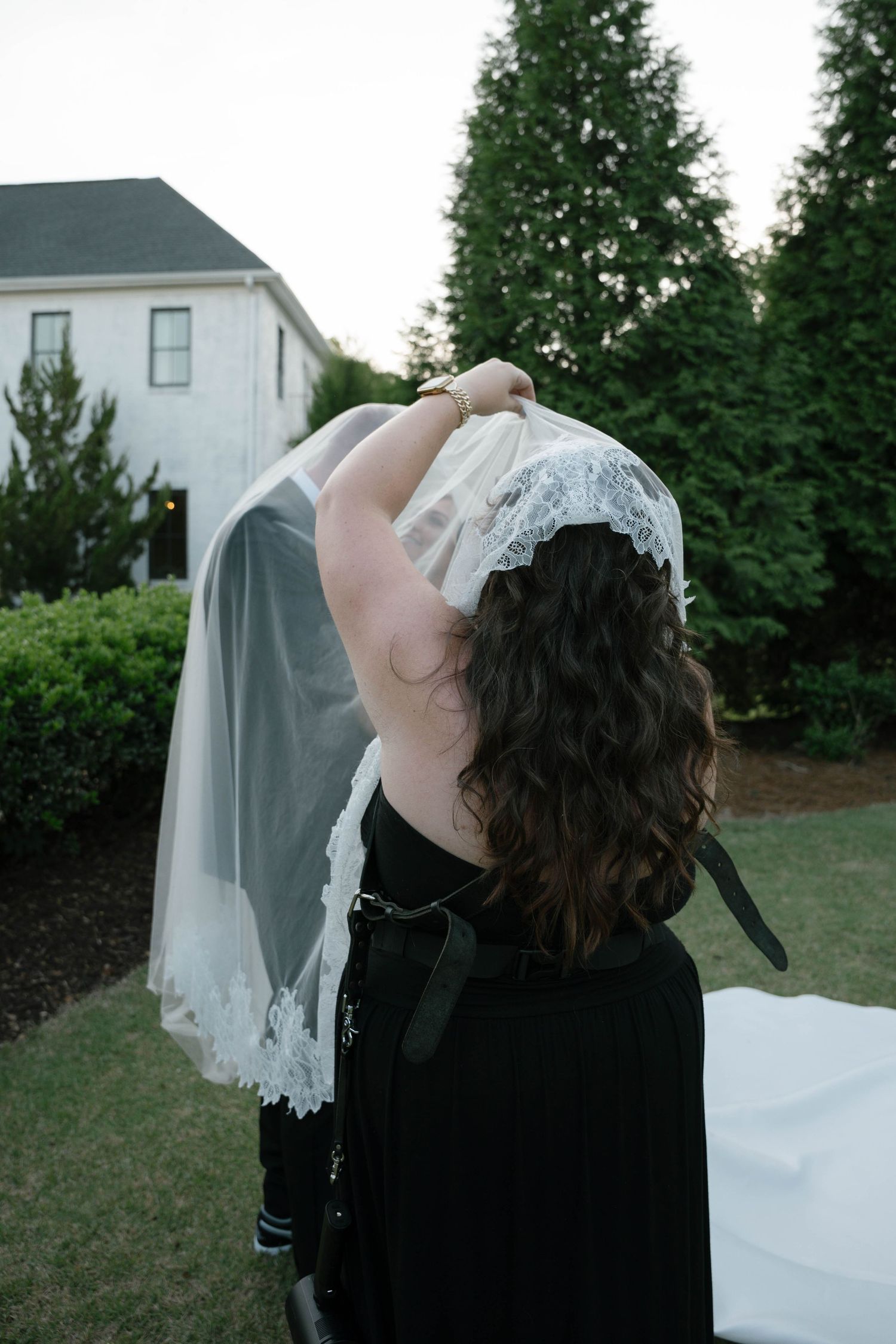 Woman in black dress holding up a white lace wedding veil outdoors near a white house and green trees.