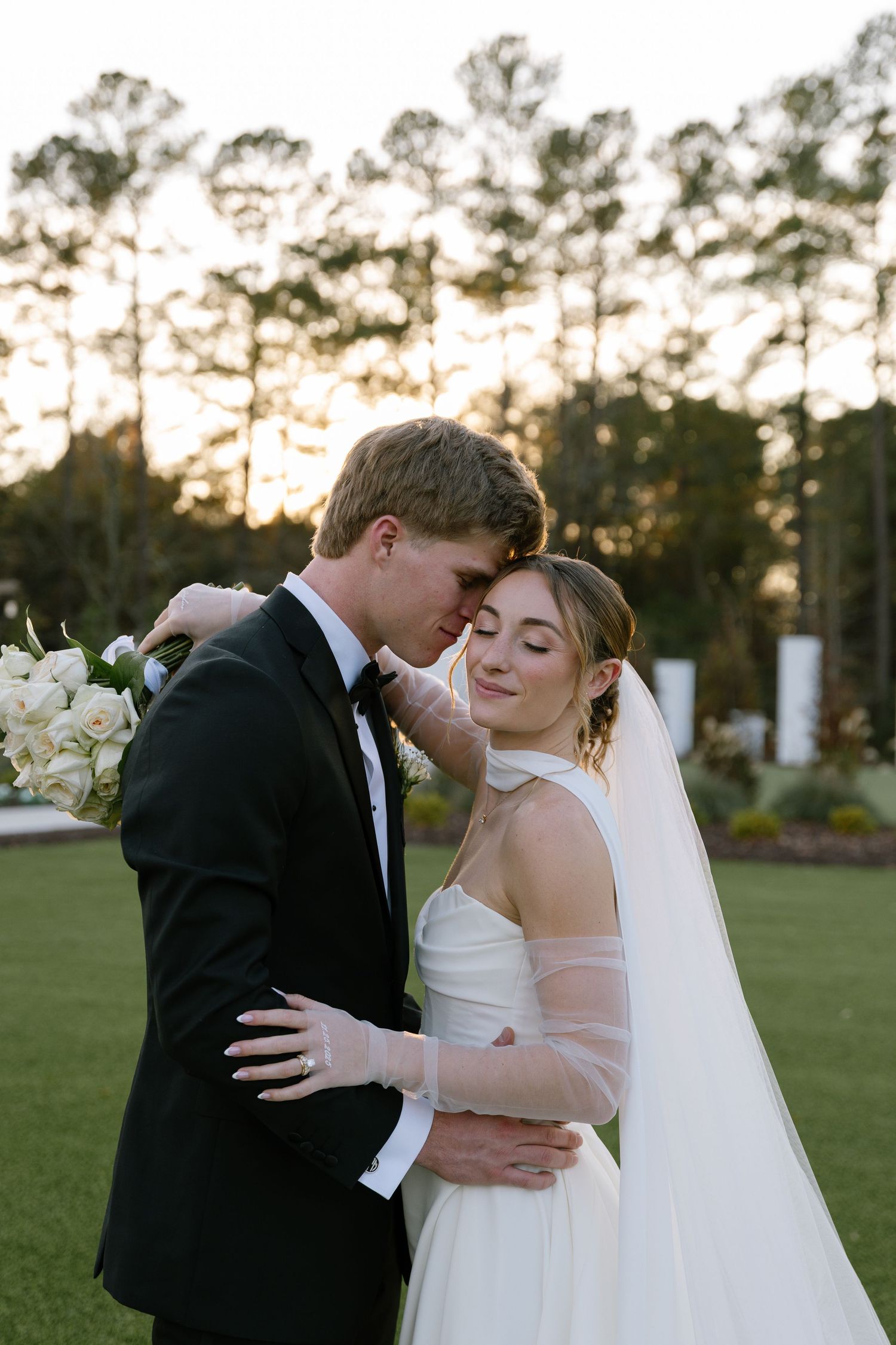 Groom in black tuxedo tenderly kisses bride's forehead outdoors at golden hour wedding portrait.