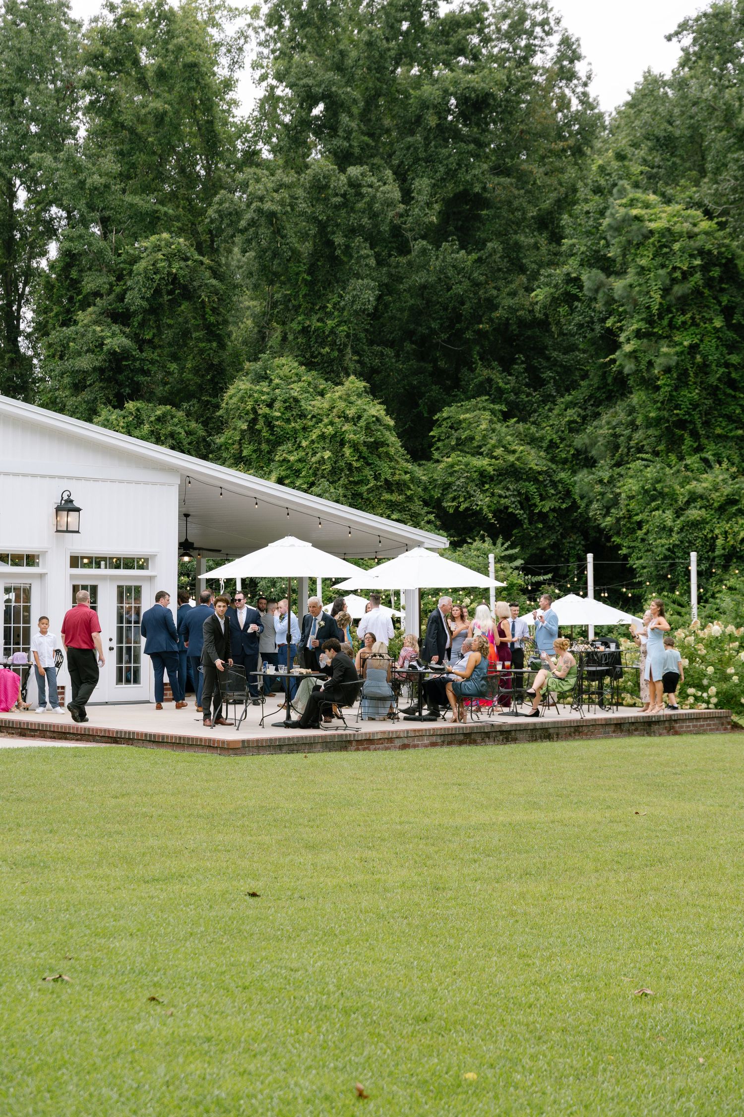 Wedding guests mingle on a white venue patio with umbrellas surrounded by lush green trees and lawn.