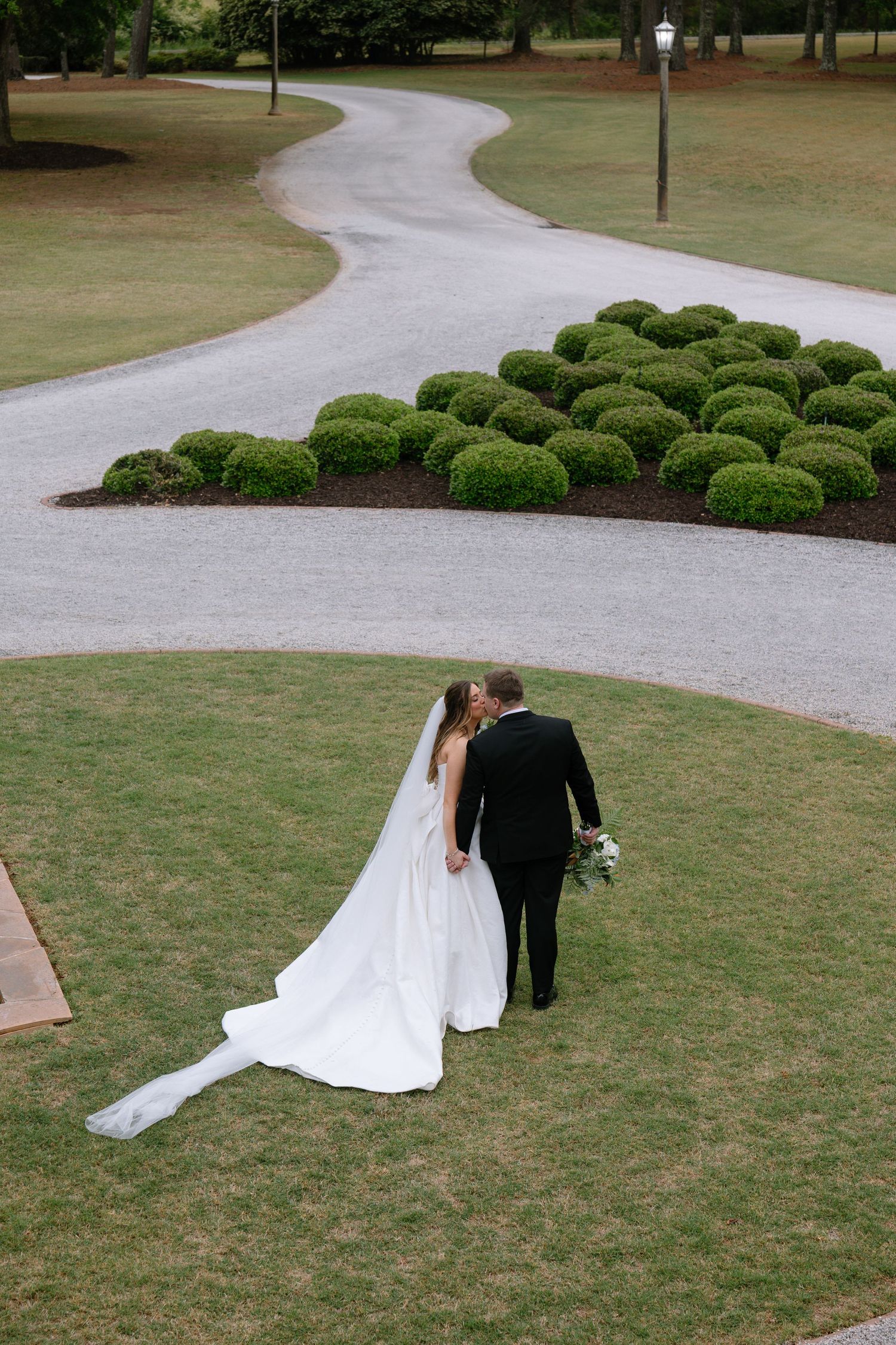 Bride and groom share a kiss on lush green lawn beside winding gravel path with manicured hedges at elegant wedding venue.