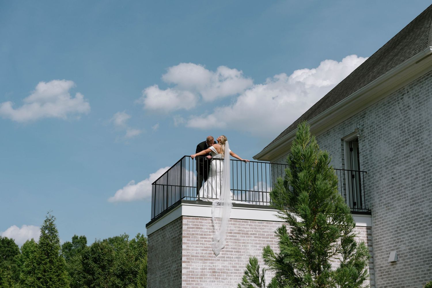 A couple embraces on a rooftop balcony of a brick building surrounded by green trees under a blue sky.