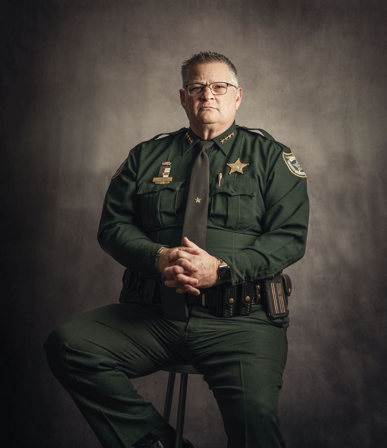 A stern law enforcement officer in green sheriff uniform sits posed for a formal portrait against a dark studio background.