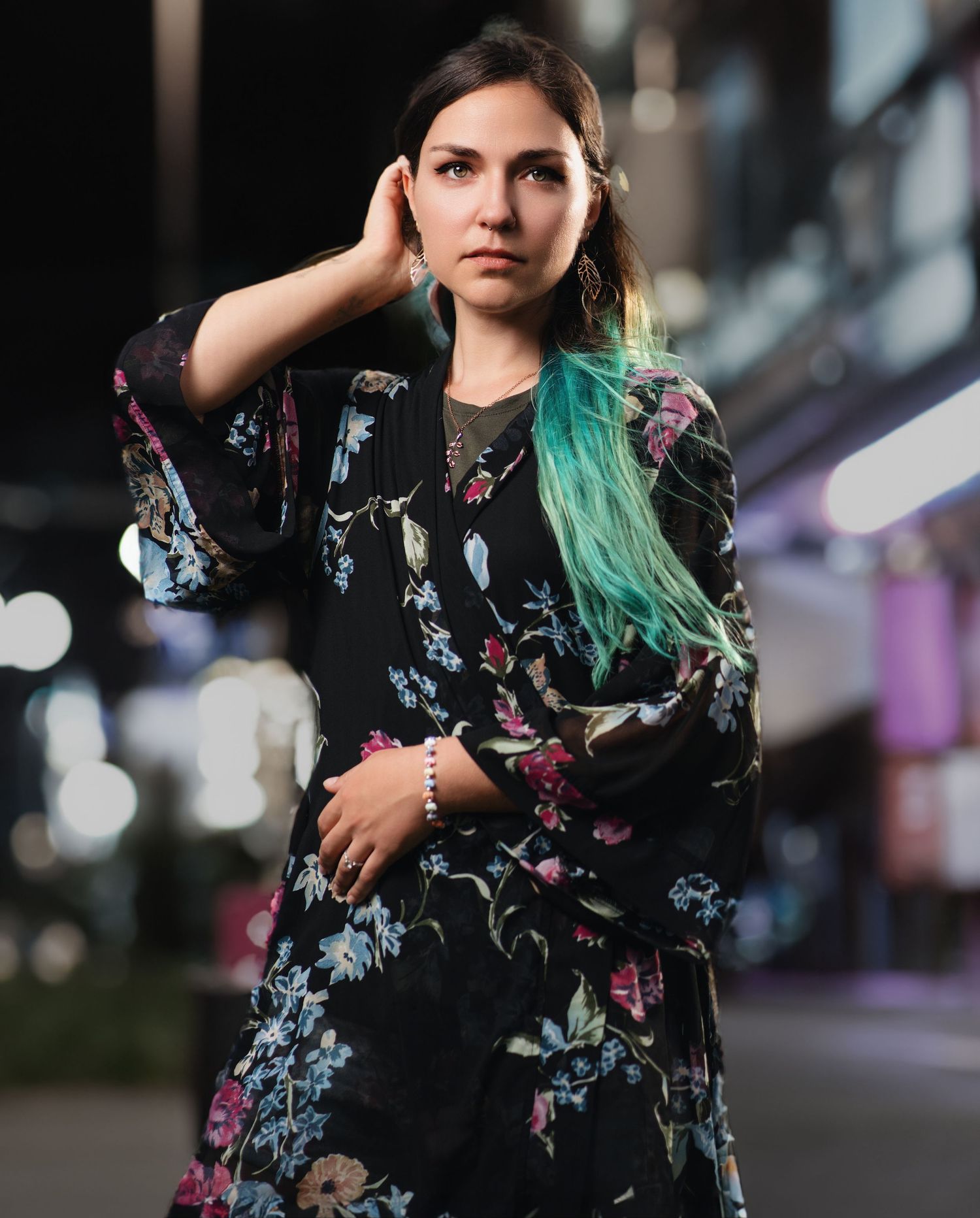 Woman with teal hair wearing a black floral kimono poses outdoors at night with city lights in background.