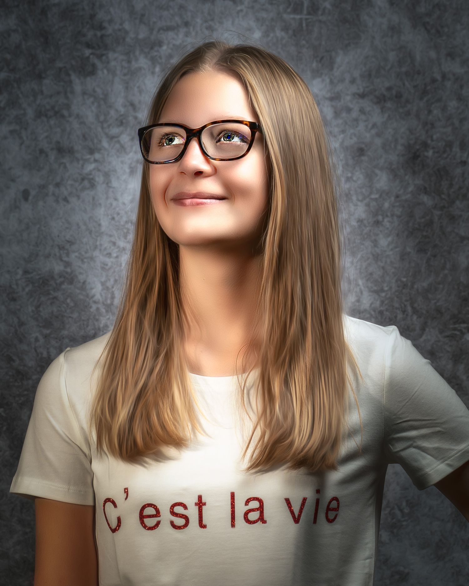 Young woman in glasses wearing a white C'est la vie t-shirt, smiling confidently against a gray background.