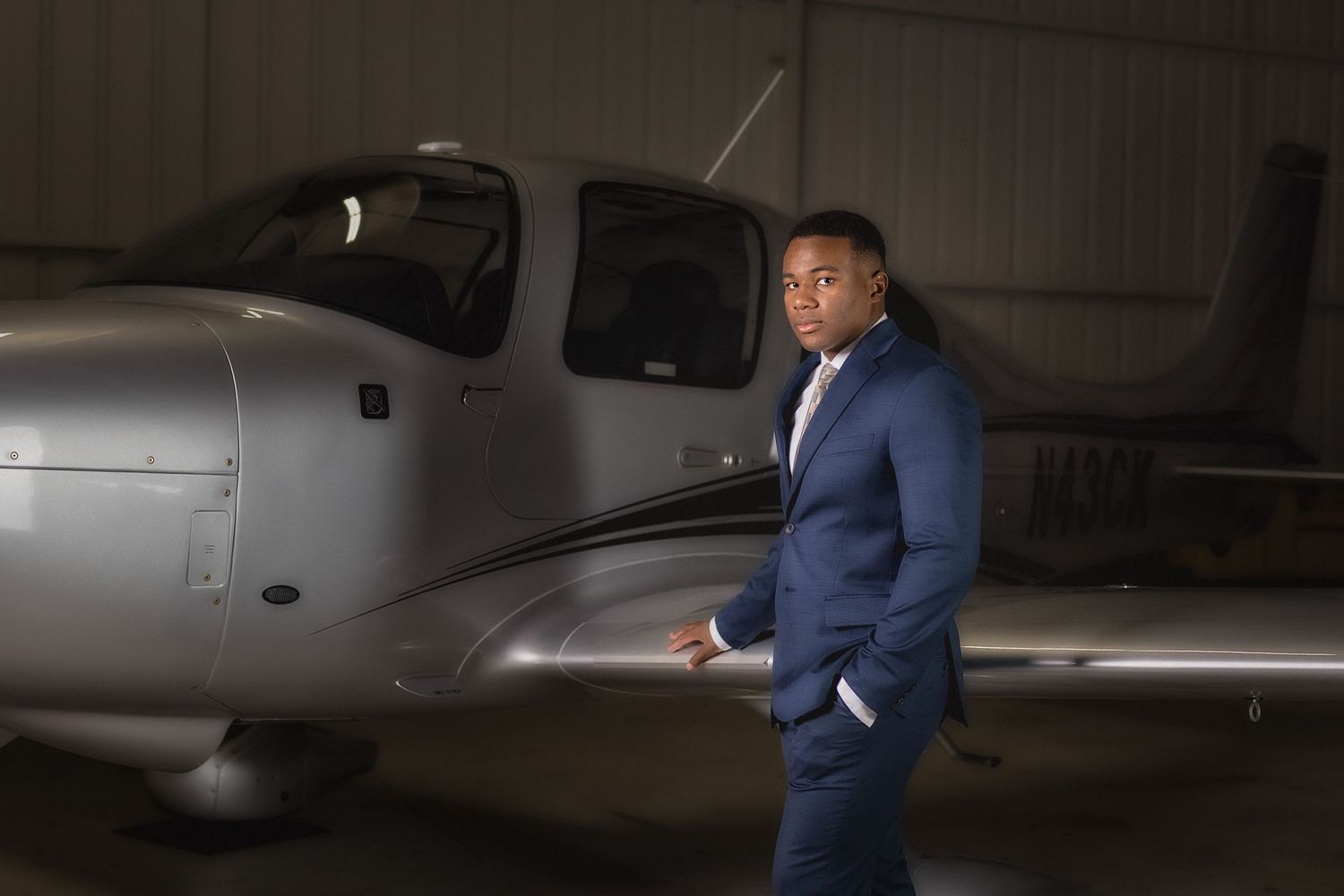 A well-dressed man in a navy blue suit stands confidently in a hangar beside a sleek silver private aircraft.