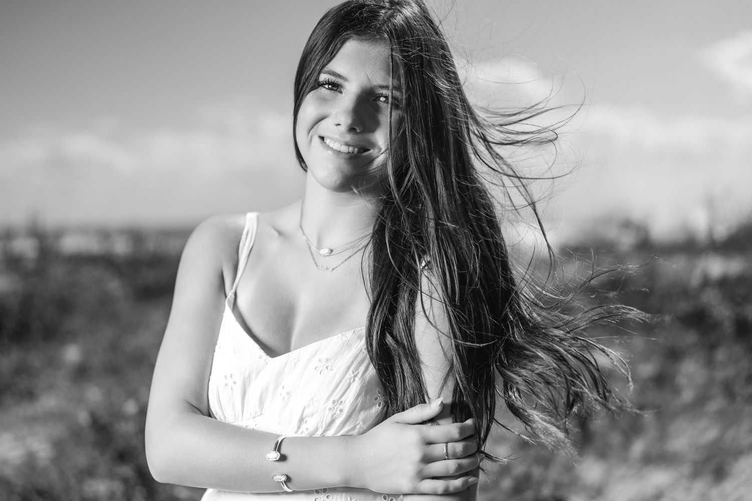 Black and white portrait of a smiling young woman with long windswept hair outdoors.