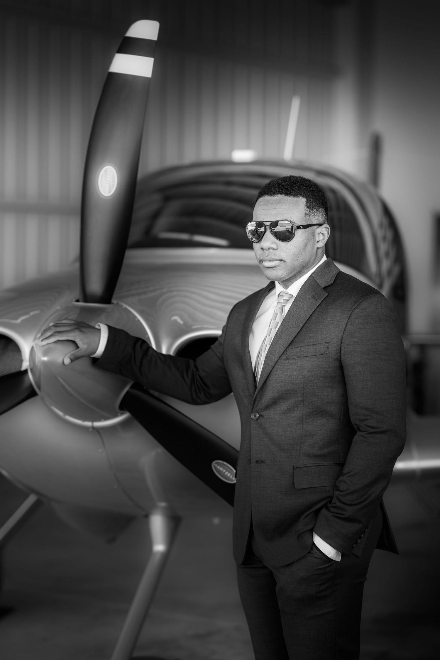 A confident man in a suit and sunglasses poses beside a propeller aircraft in a hangar, black and white photo.